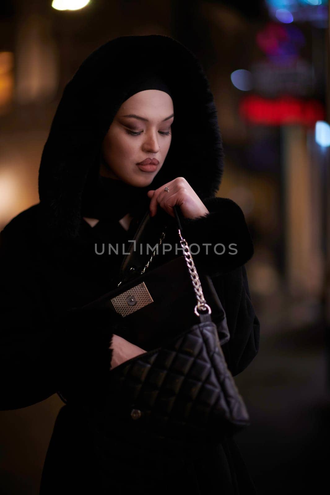 Young Muslim woman walking on urban city street on a cold winter night wearing hijab scarf veil a fashionable coat with bokeh city light in the background.