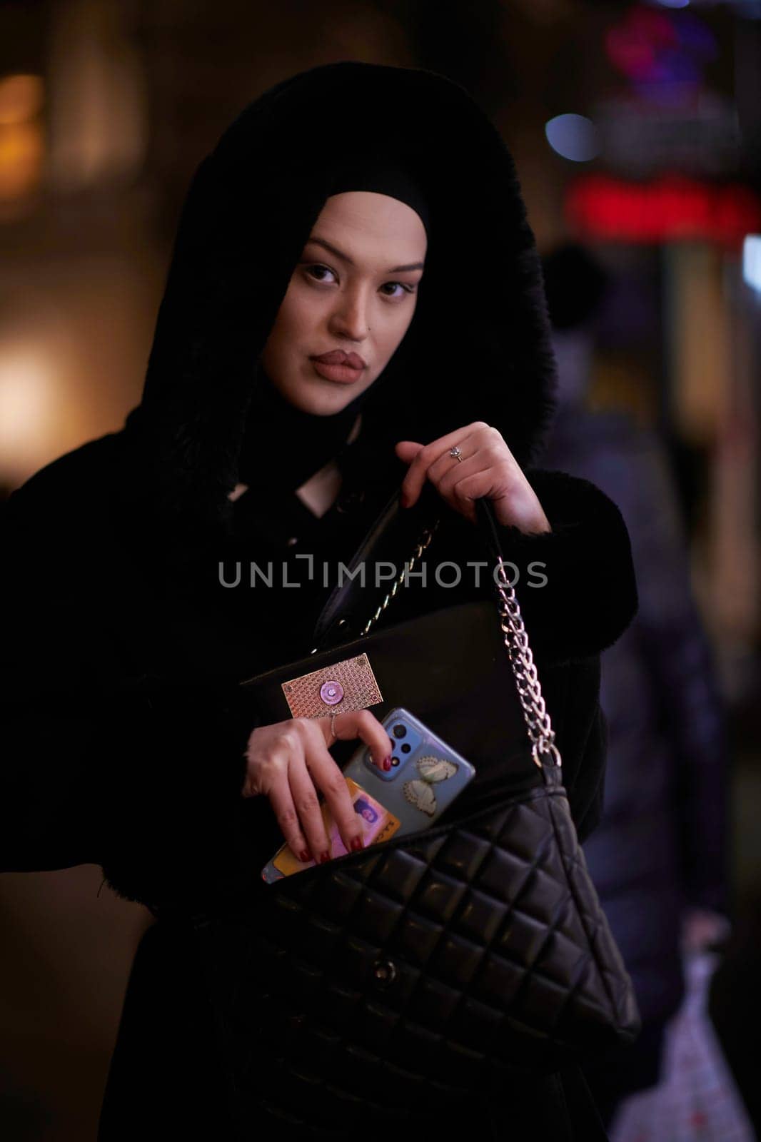 Young Muslim woman walking on urban city street on a cold winter night wearing hijab scarf veil a fashionable coat with bokeh city light in the background.