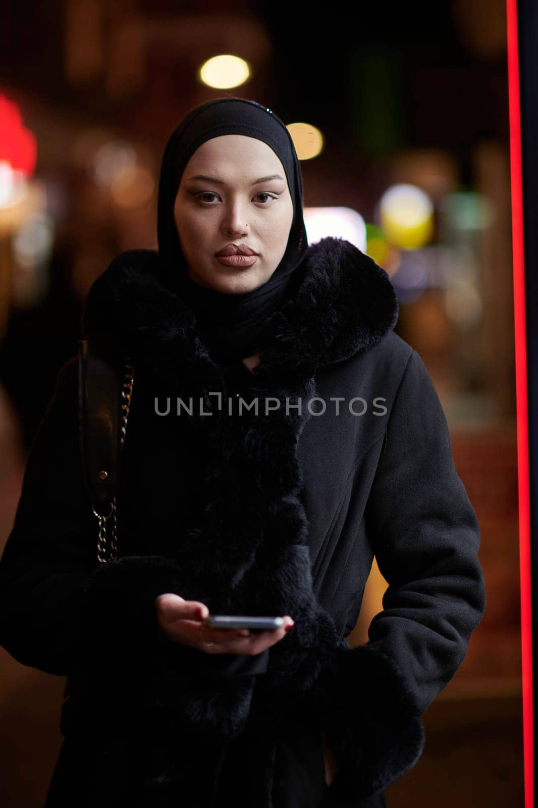 Young Muslim woman walking on urban city street on a cold winter night wearing hijab scarf veil a fashionable coat with bokeh city light in the background.