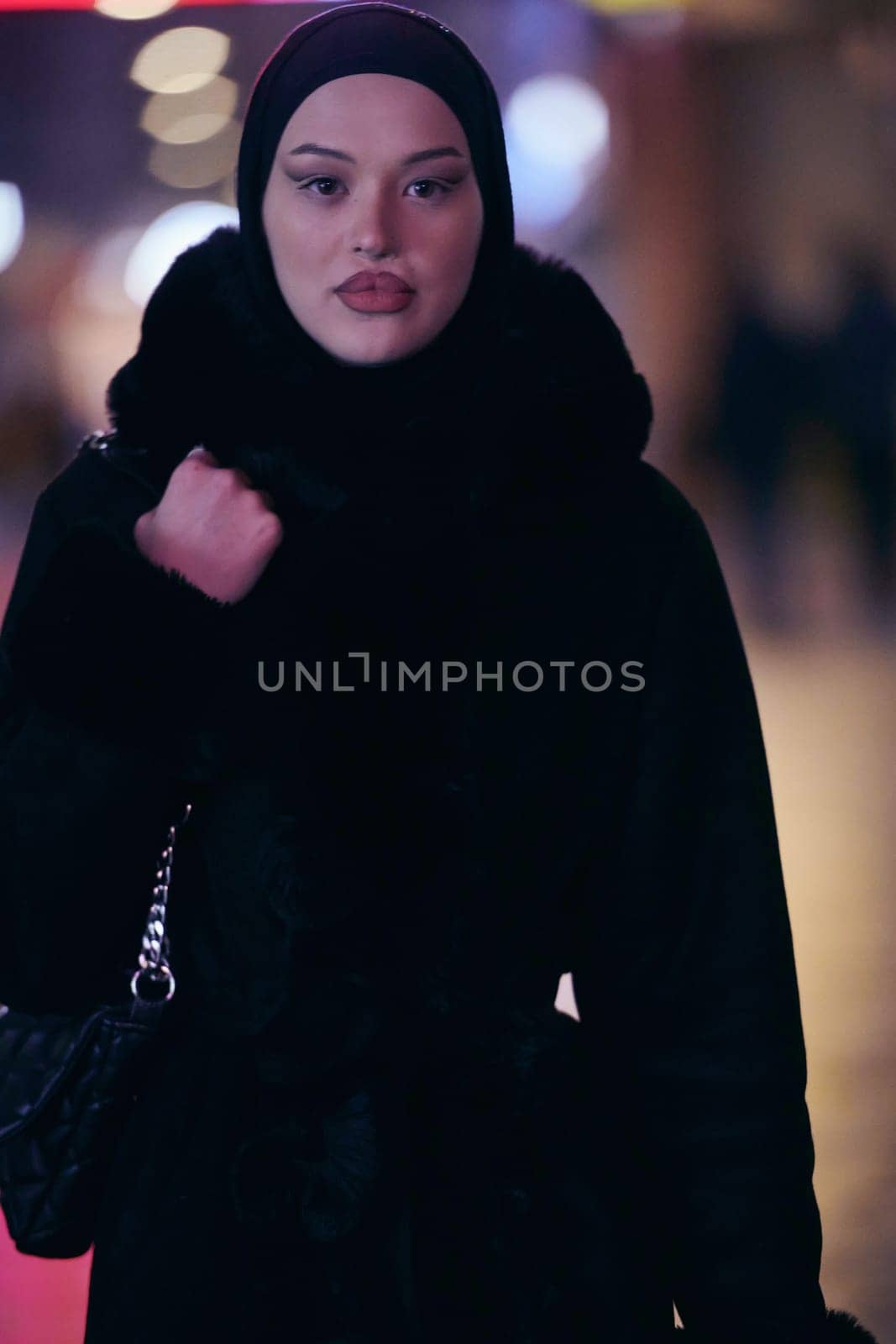 Young Muslim woman walking on urban city street on a cold winter night wearing hijab scarf veil a fashionable coat with bokeh city light in the background.