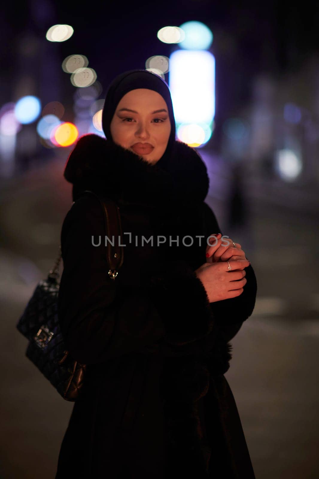 Young Muslim woman walking on urban city street on a cold winter night wearing hijab scarf veil a fashionable coat with bokeh city light in the background.