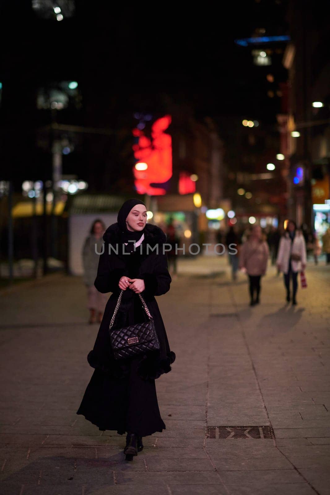 Young Muslim woman walking on urban city street on a cold winter night wearing hijab scarf veil a fashionable coat with bokeh city light in the background.