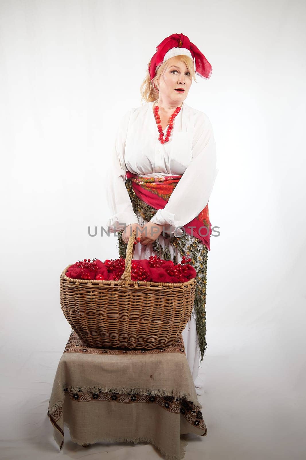 Cheerful funny adult mature woman solokha in a red knitted shawl. Female model in clothes of national ethnic Slavic style. Stylized Ukrainian, Belarusian or Russian woman poses in comic photo shoot