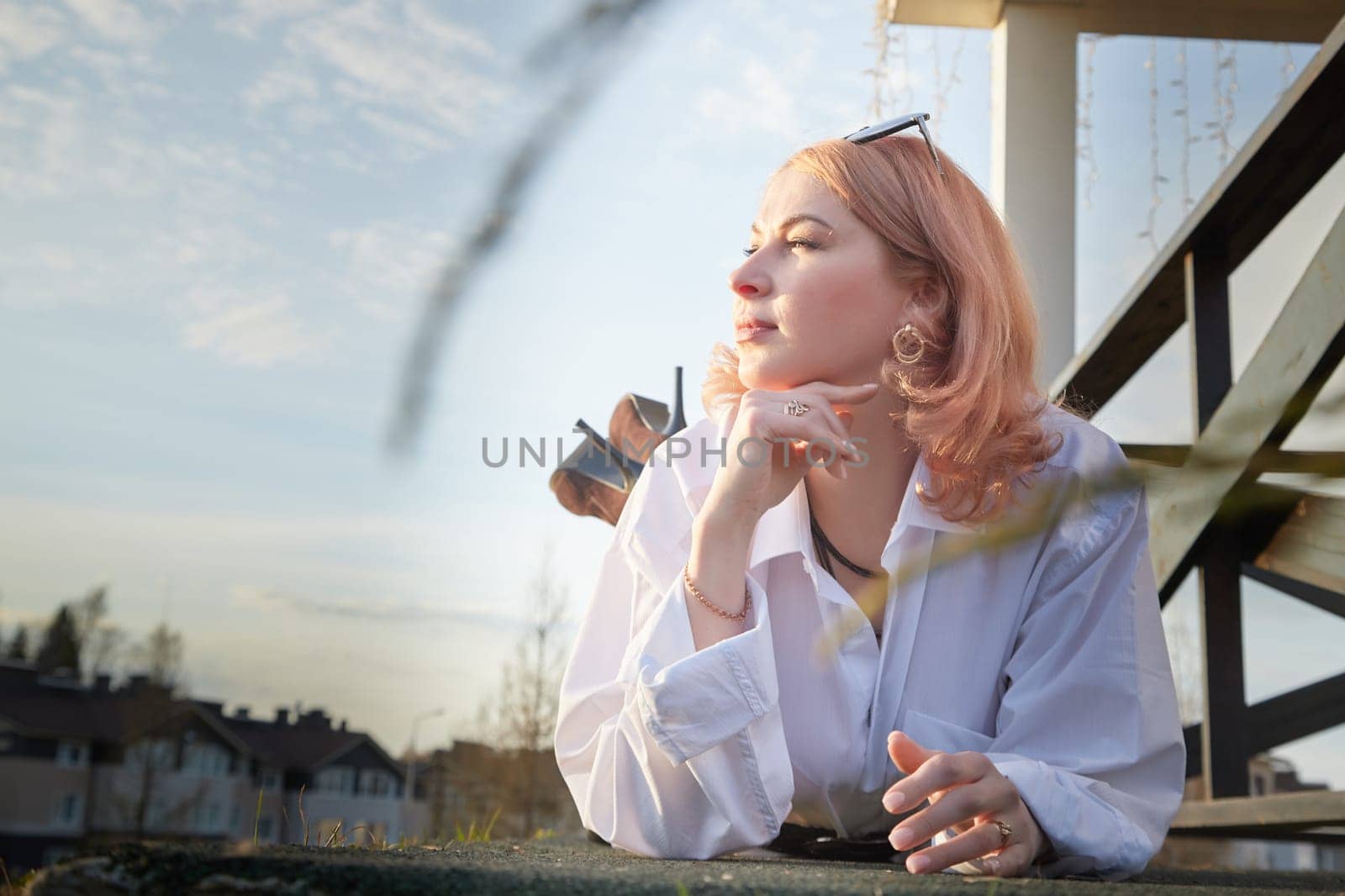 Portrait of Beautiful girl with red hair in white shirt in open wooden pavillion in village or small town. Young slender woman and sky with clouds on background on autumn, spring or summer evening