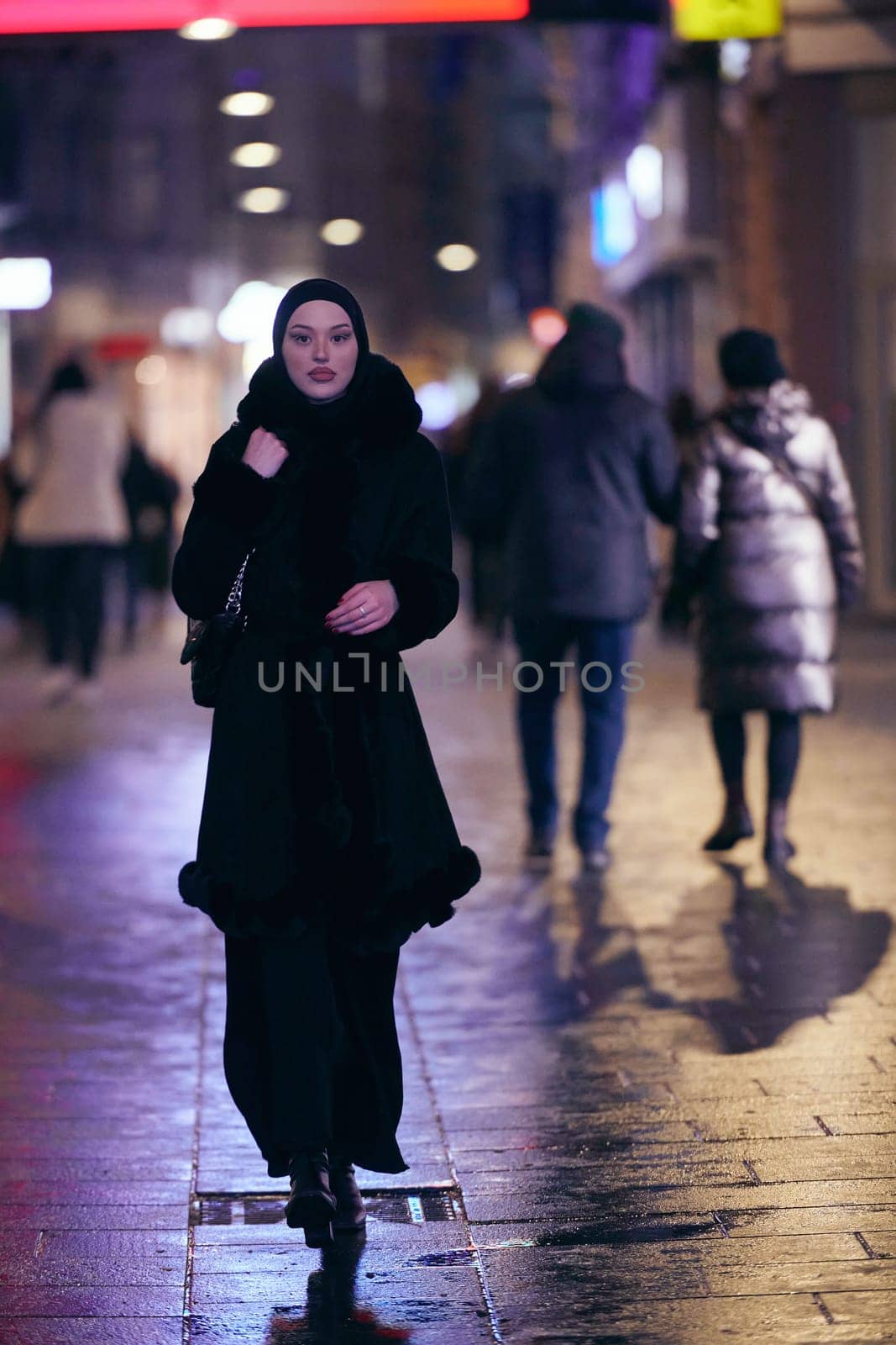 Young Muslim woman walking on urban city street on a cold winter night wearing hijab scarf veil a fashionable coat with bokeh city light in the background.