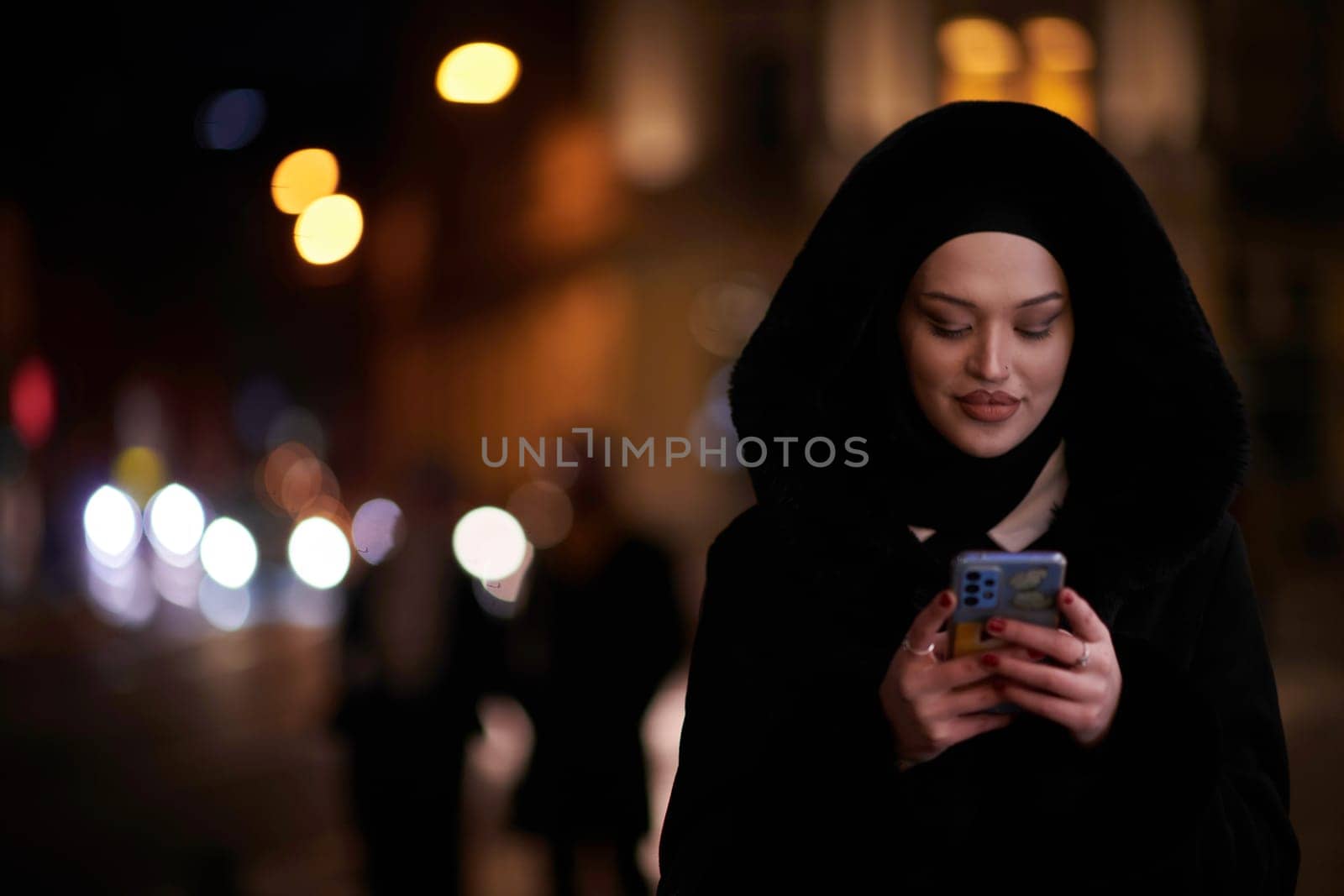 Young Muslim woman walking on urban city street on a cold winter night wearing hijab scarf veil a fashionable coat with bokeh city light in the background.