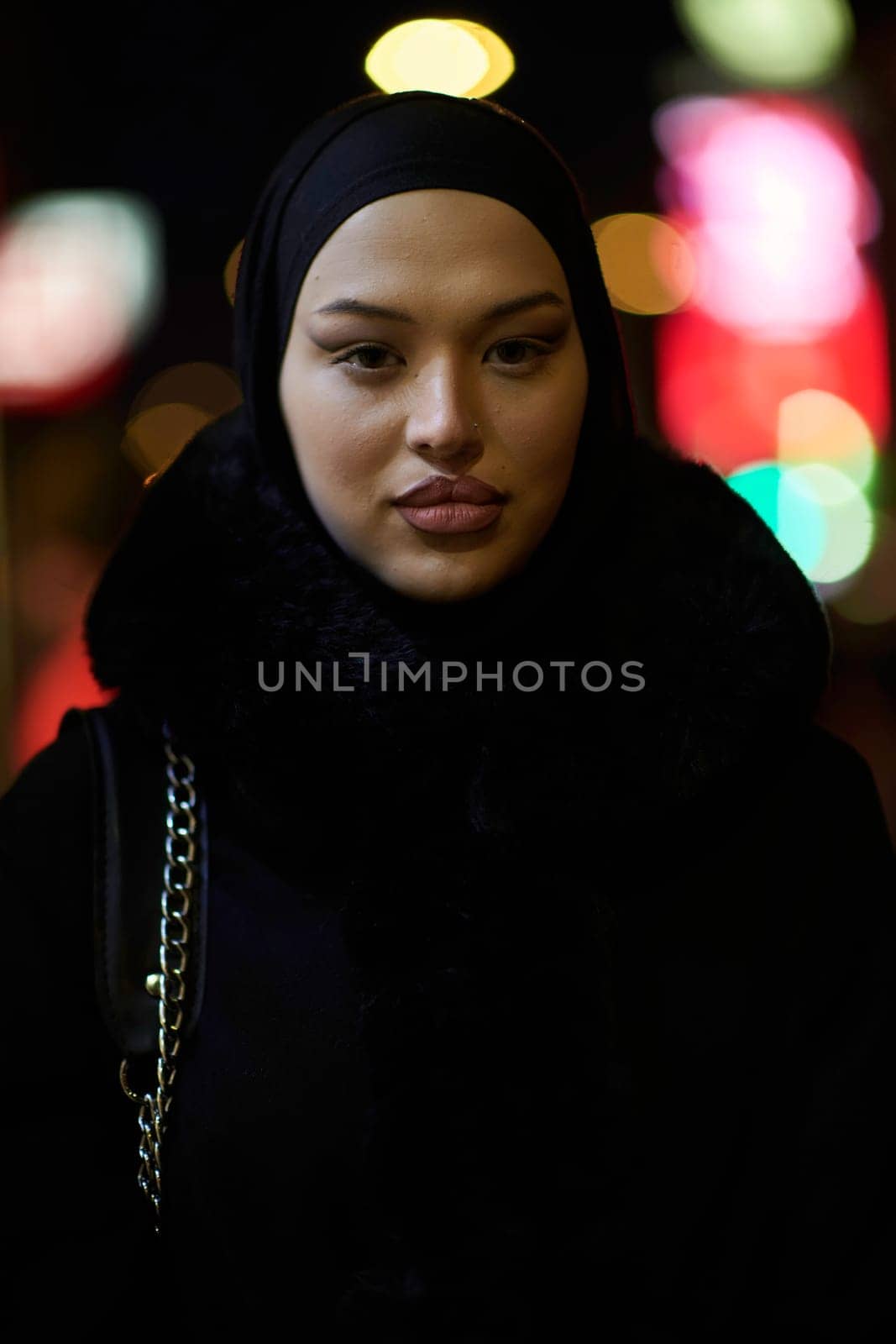 Young Muslim woman walking on urban city street on a cold winter night wearing hijab scarf veil a fashionable coat with bokeh city light in the background.