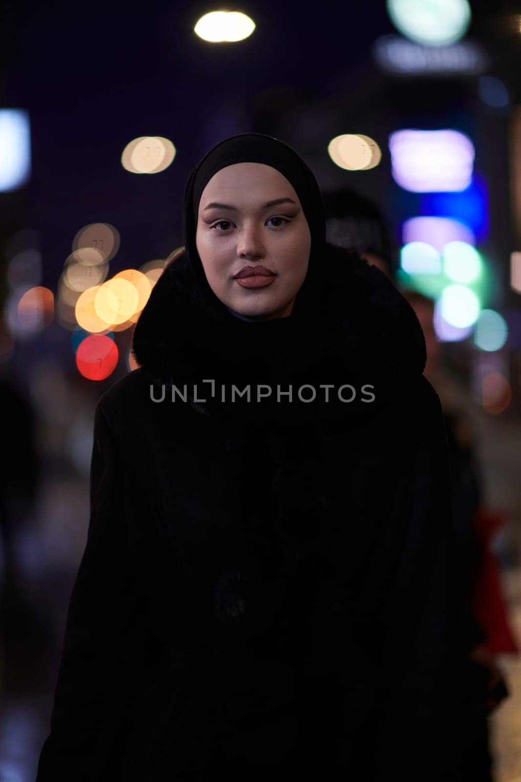 Young Muslim woman walking on urban city street on a cold winter night wearing hijab scarf veil a fashionable coat with bokeh city light in the background.