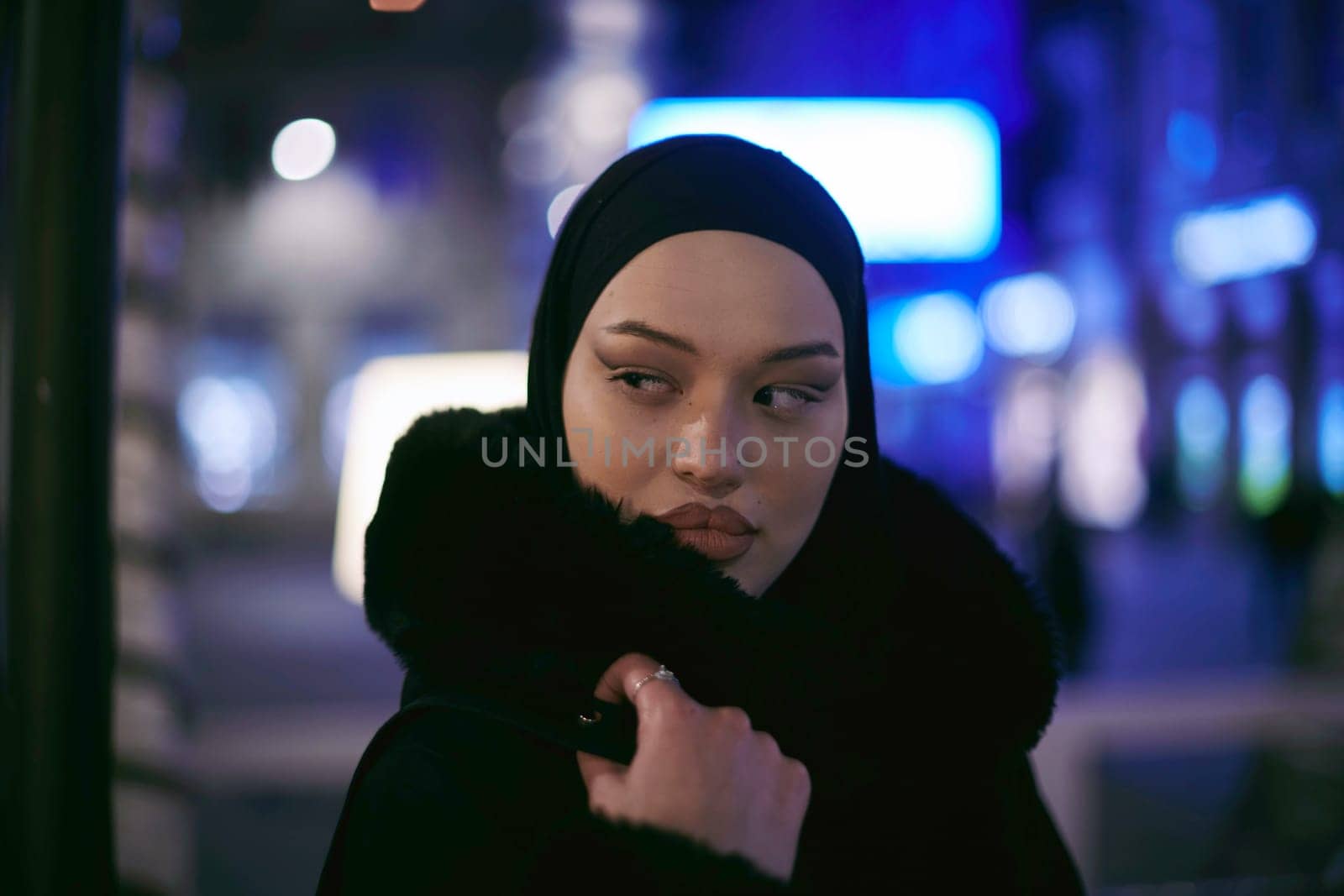 Young Muslim woman walking on urban city street on a cold winter night wearing hijab scarf veil a fashionable coat with bokeh city light in the background.