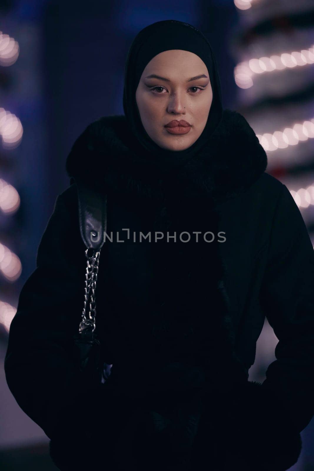 Young Muslim woman walking on urban city street on a cold winter night wearing hijab scarf veil a fashionable coat with bokeh city light in the background.