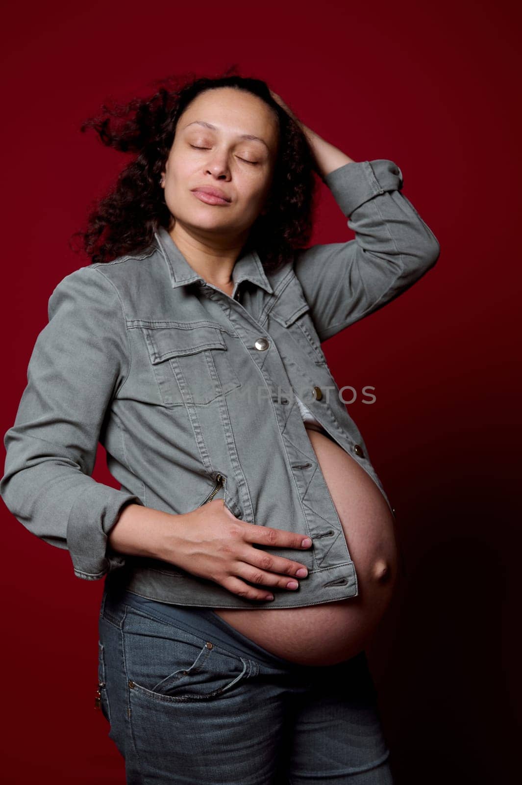 Beautiful multi-ethnic young pregnant woman, expectant mother with naked belly, wearing stylish casual denim blazer and jeans, posing over isolated red color studio background