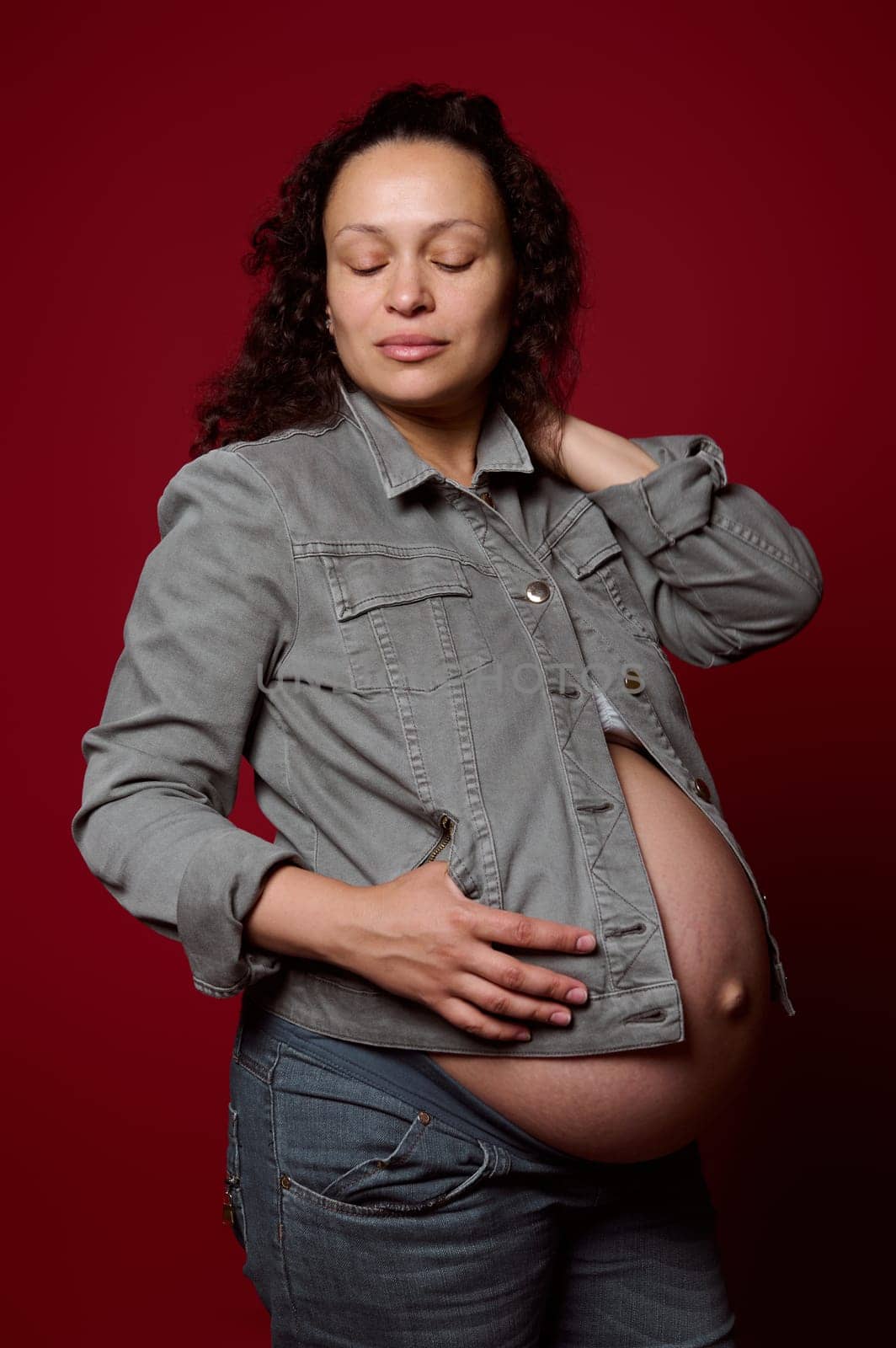 Multi-ethnic pregnant woman, expectant mother in casual denim clothes, posing with her eyes closed over red isolated studio background. Pregnancy 36 week. Childbearing. Maternity lifestyle