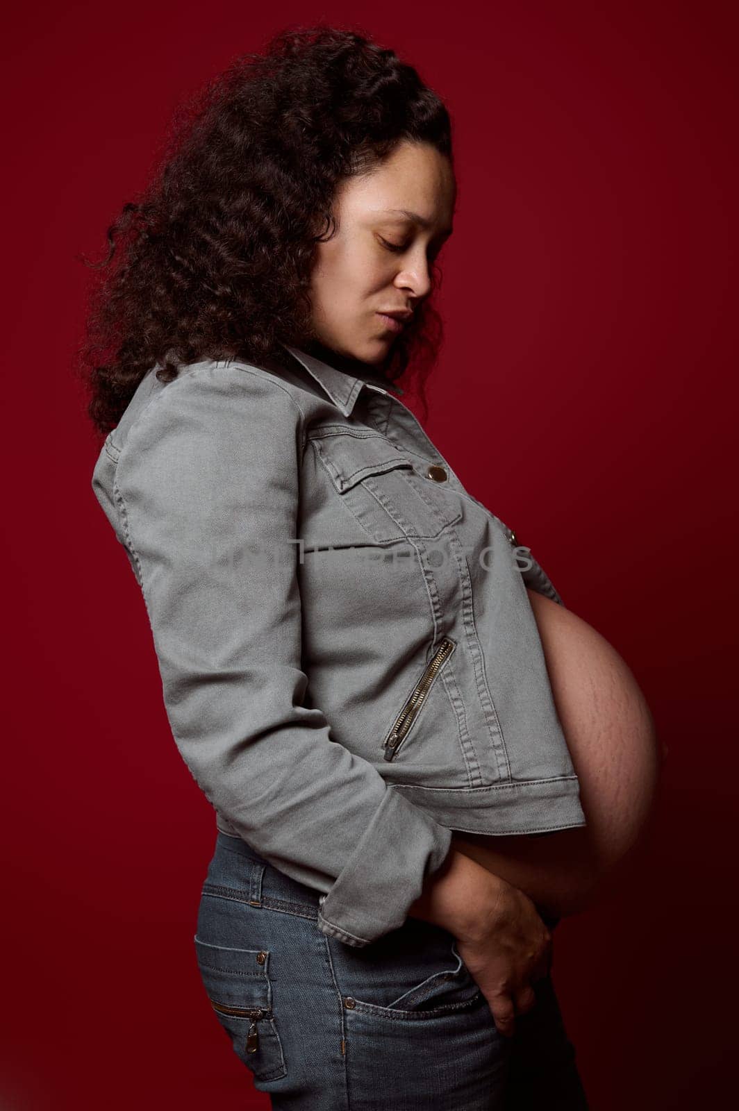 Side portrait of pregnant woman in casual denim clothes, posing with her eyes closed over red isolated studio background by artgf