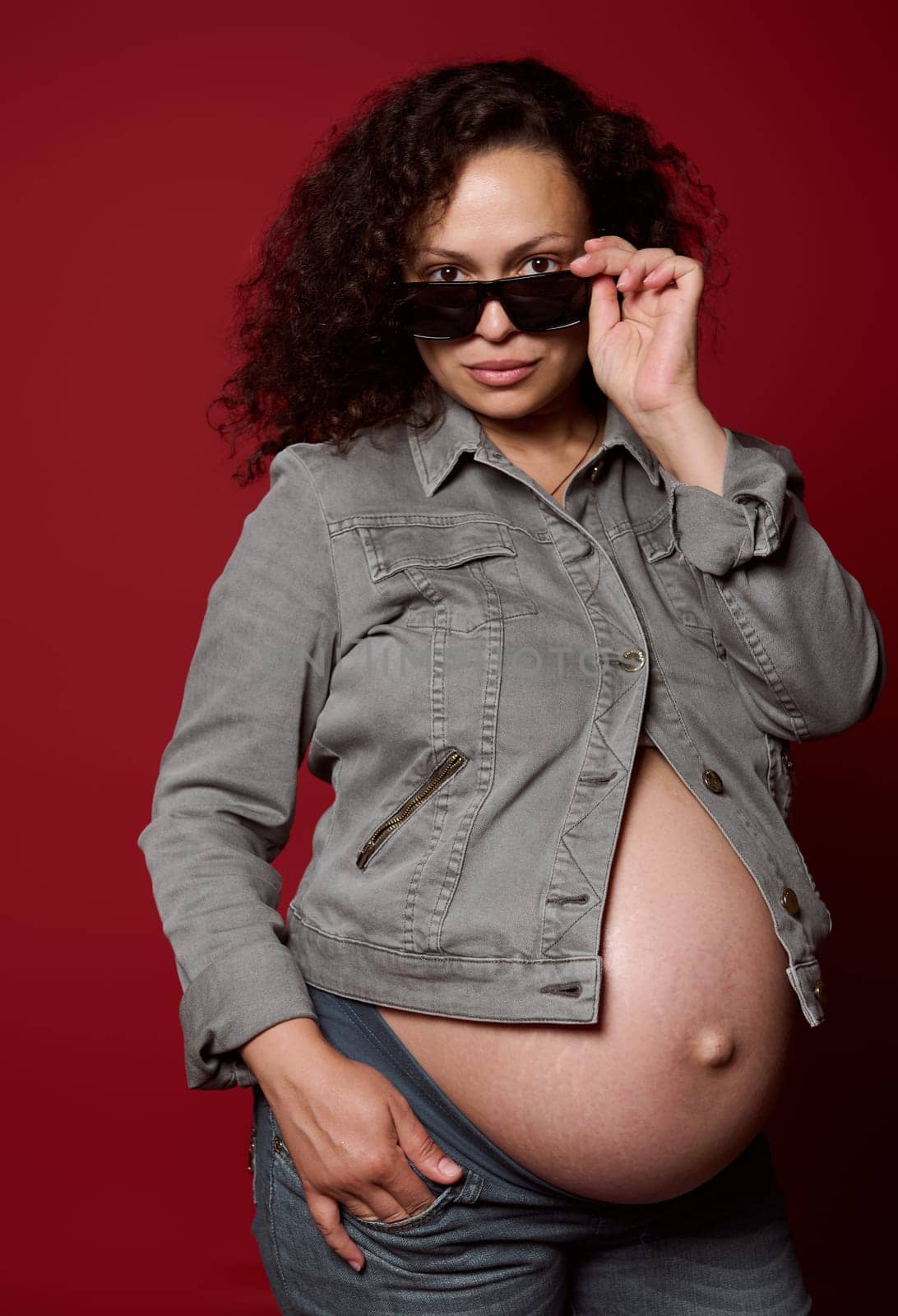 Pregnant woman dressed in casual jeans clothes, looking at camera, posing with naked belly, isolated studio background by artgf