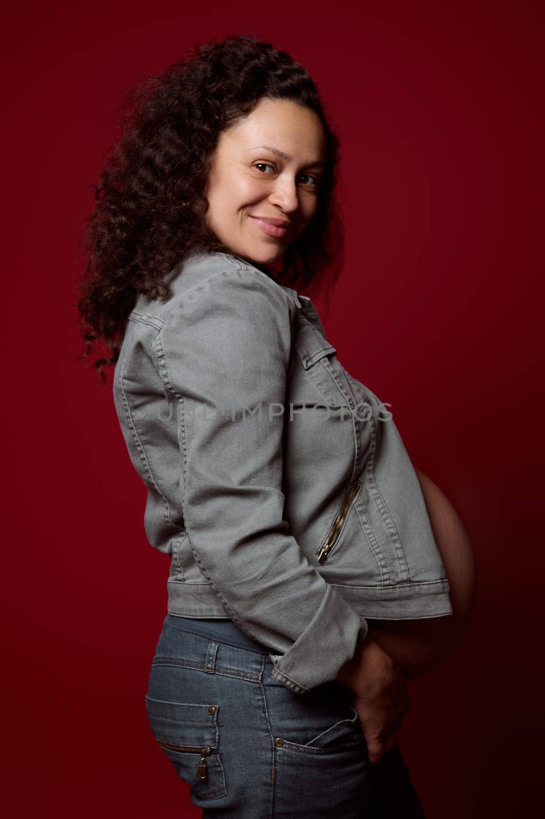 Advertising studio shot of a beautiful curly haired pregnant woman in casual jeans, smiles looking at camera, isolated over red studio background. Pregnancy and maternity fashion, people, lifestyle