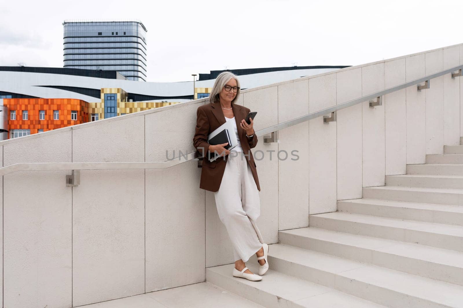 a well-groomed middle-aged woman in glasses dressed in a brown jacket is waiting for a business partner on the street.