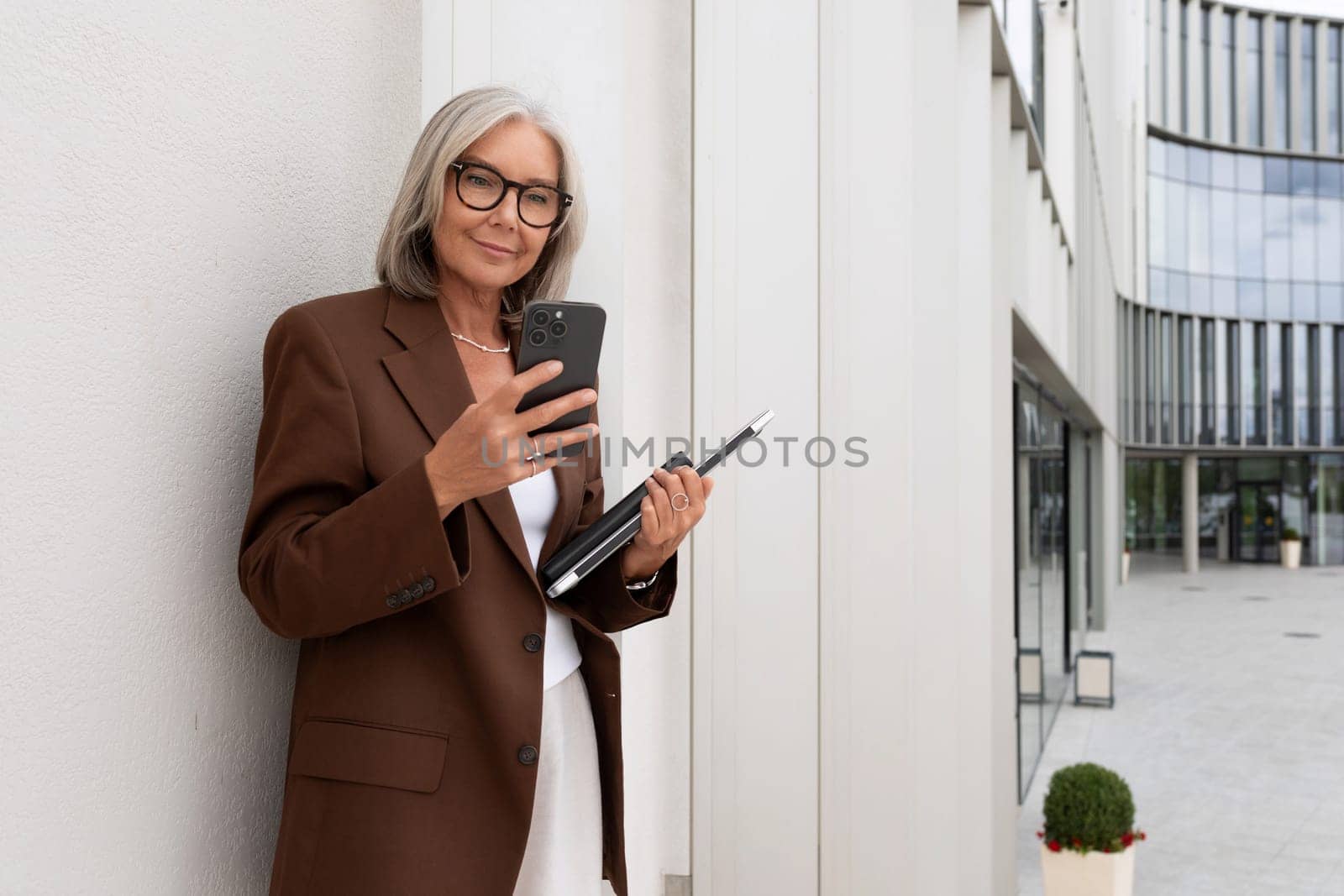a gray-haired middle-aged woman with a bob hairstyle dressed in a jacket and trousers walks around the city against the backdrop of a business center and offices by TRMK