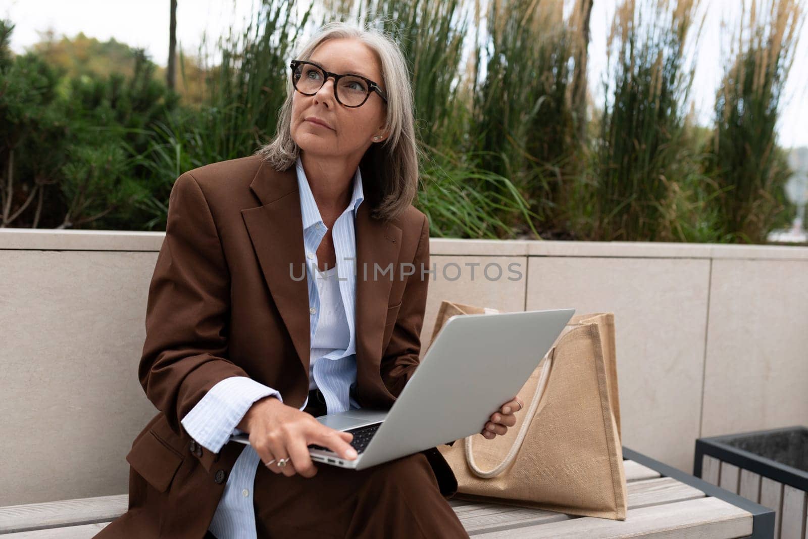 well-groomed charming entrepreneur woman of mature years works remotely sitting on a bench with a laptop by TRMK