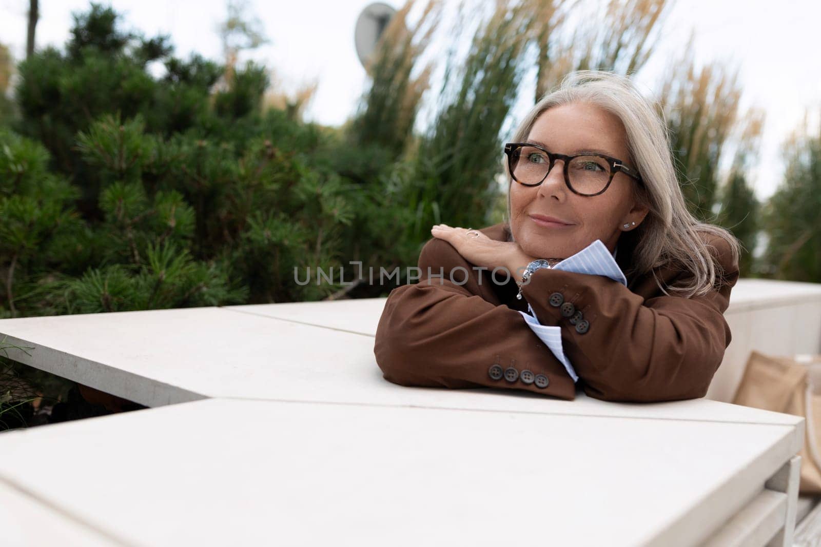 a mature woman with gray hair in a brown jacket is sitting at a table on the terrace of a cafe waiting for a partner by TRMK