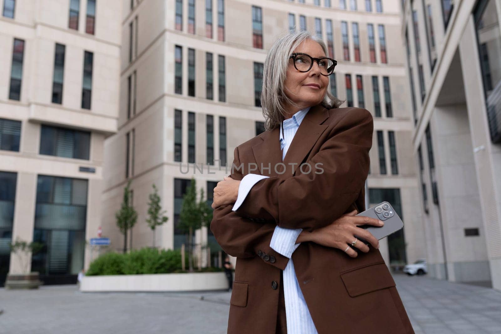portrait of a confident successful 60 year old gray-haired business lady in glasses wearing a brown jacket on the street.