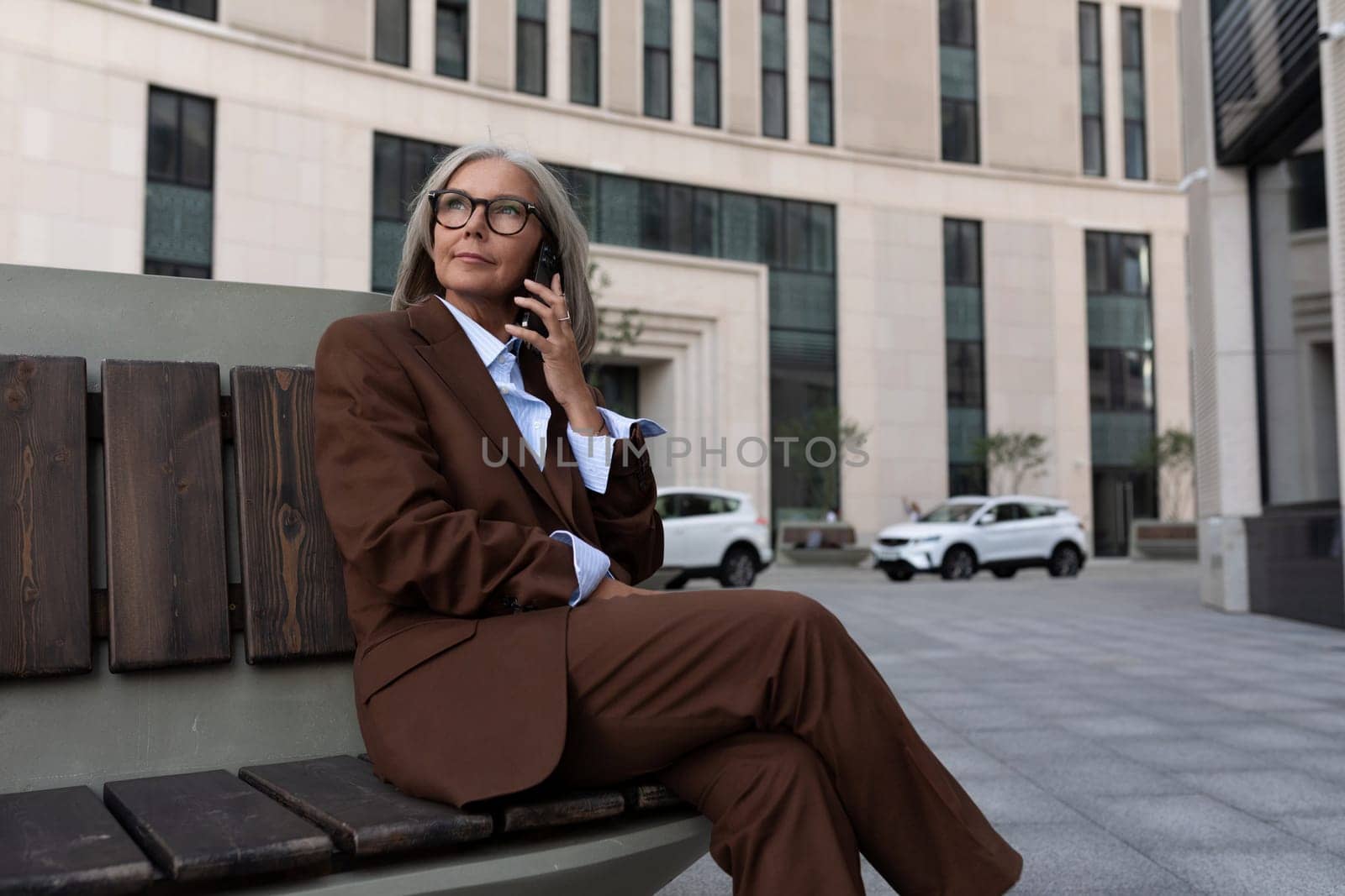 portrait of a confident successful 60 year old gray-haired business lady in glasses wearing a brown jacket on the street by TRMK