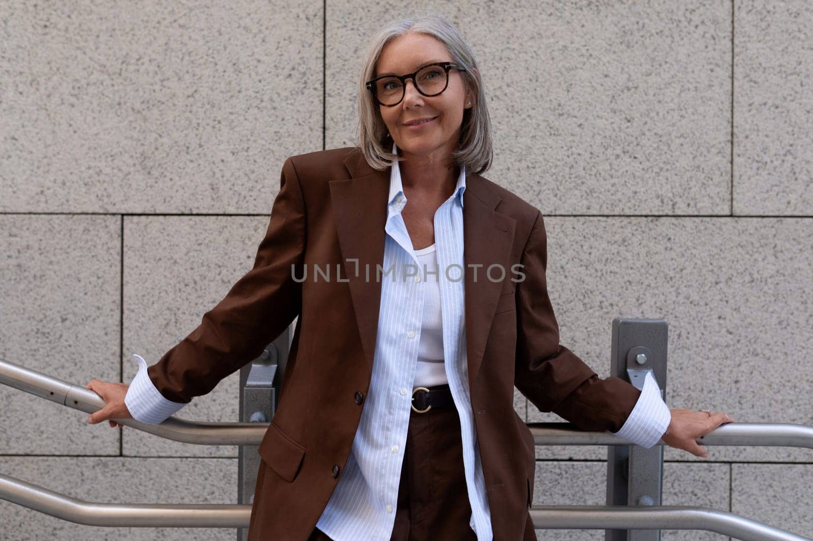 portrait of a slender cute gray-haired entrepreneur woman of mature years in a brown suit on the background of the building by TRMK