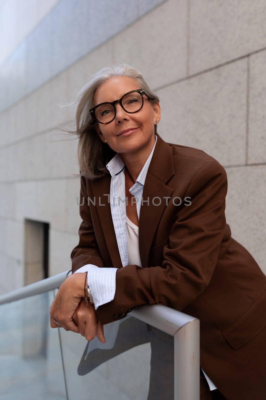 business portrait of a confident successful 60 year old gray-haired lady in glasses wearing a brown jacket.