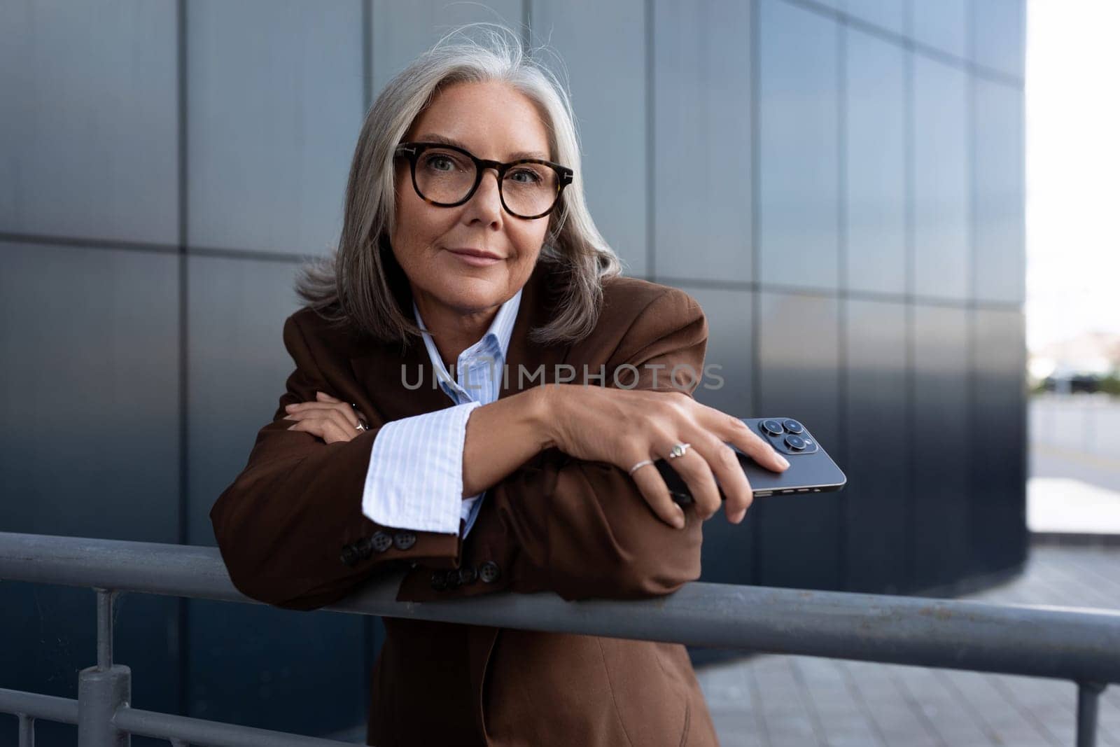 charming gray-haired well-groomed middle-aged woman in glasses dressed in a brown jacket on the street by TRMK