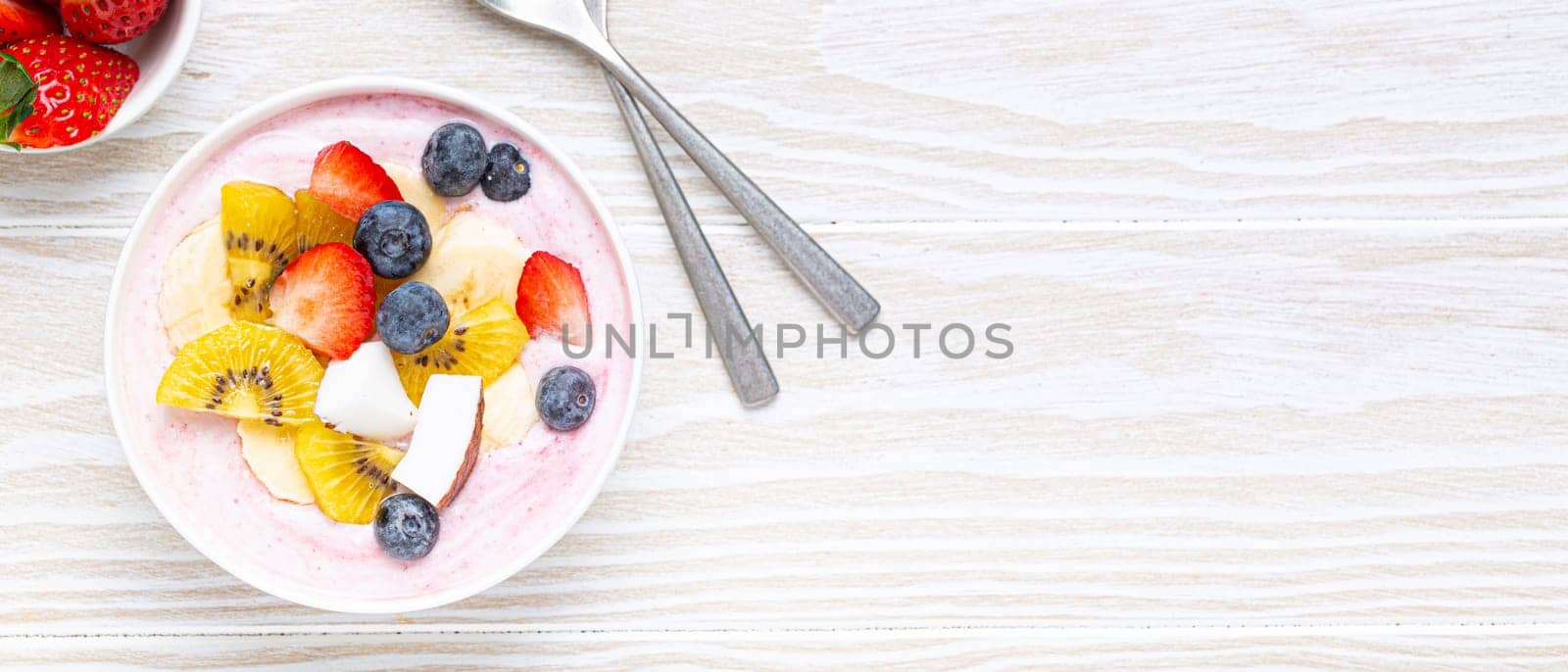 Healthy breakfast or dessert yogurt bowl with fresh banana, strawberry, blueberry, cocos, kiwi top view on rustic wooden white background with spoon. Space for text