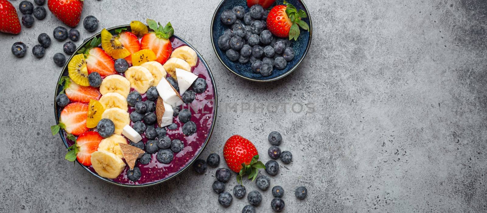 Healthy summer acai smoothie bowl with chia seeds, fresh banana, strawberry, blueberry, cocos, kiwi top view, rustic concrete background with spoon. Copy space by its_al_dente