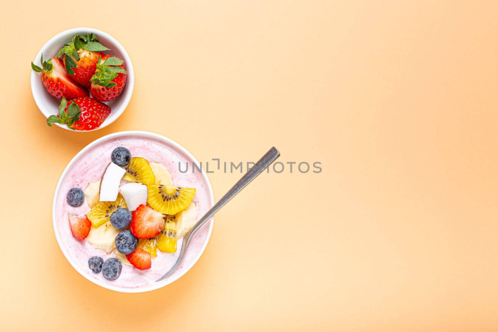 Healthy breakfast or dessert yogurt bowl with fresh banana, strawberry, blueberry, cocos, kiwi top view on minimal pastel paper background with spoon. Copy space