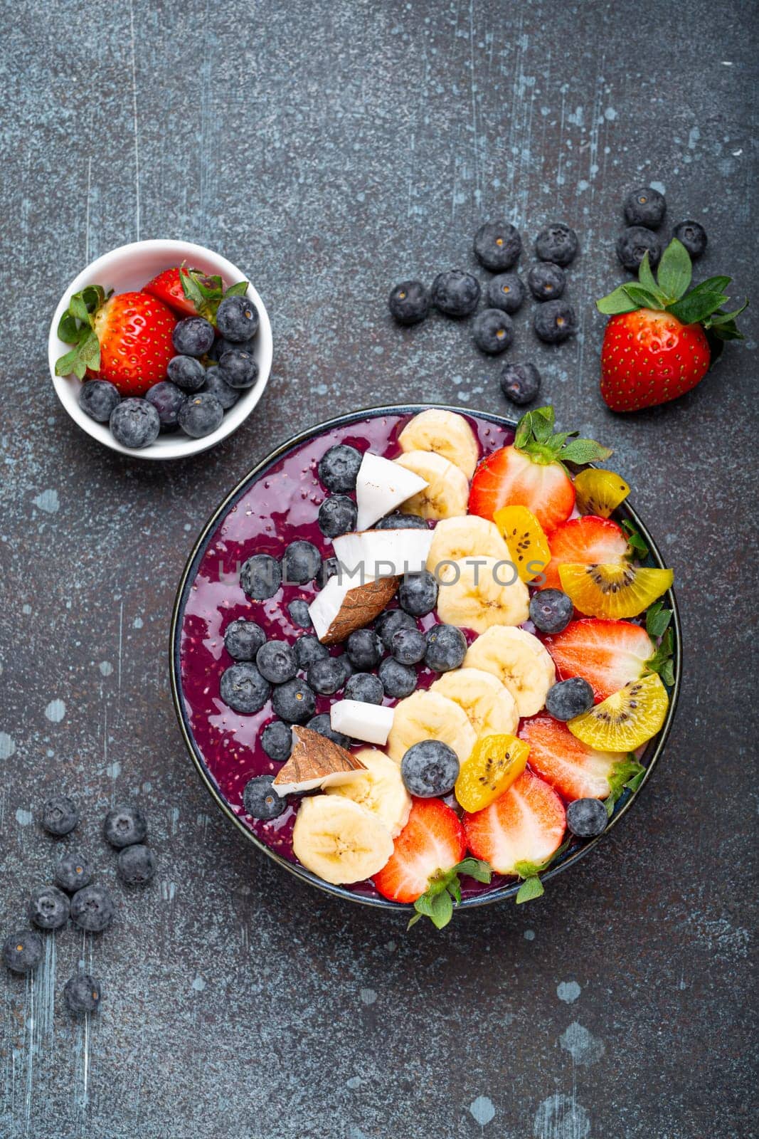 Healthy summer acai smoothie bowl with chia seeds, fresh banana, strawberry, blueberry, cocos, kiwi top view on rustic concrete background with spoon