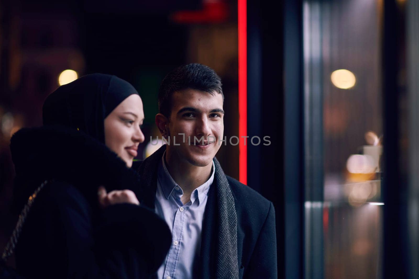 Happy multicultural business couple walking together outdoors in an urban city street at night near a jewelry shopping store window. Successful Arab businessman and European Muslim woman.