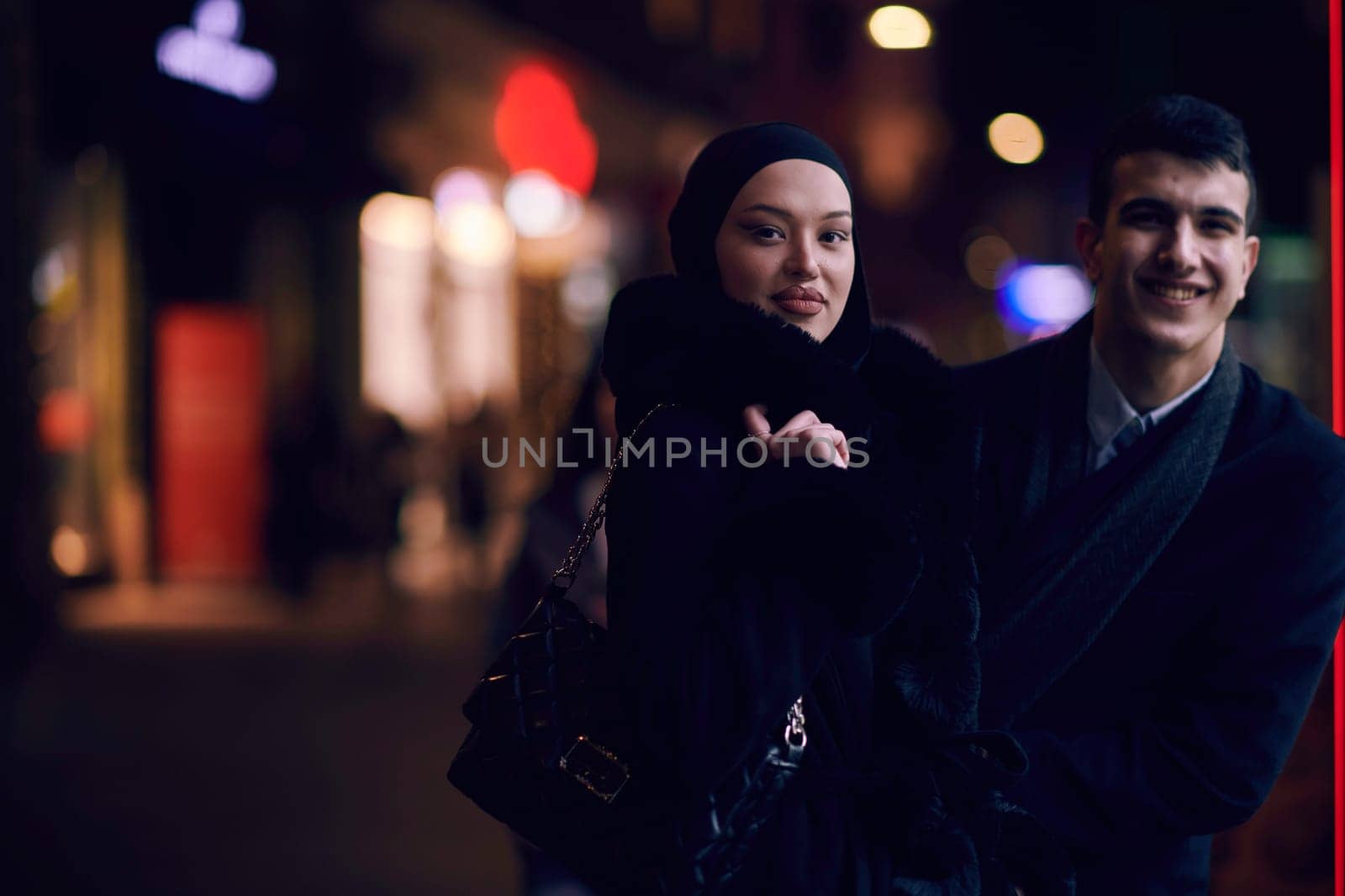 Happy multicultural business couple walking together outdoors in an urban city street at night near a jewelry shopping store window. Successful Arab businessman and European Muslim woman.