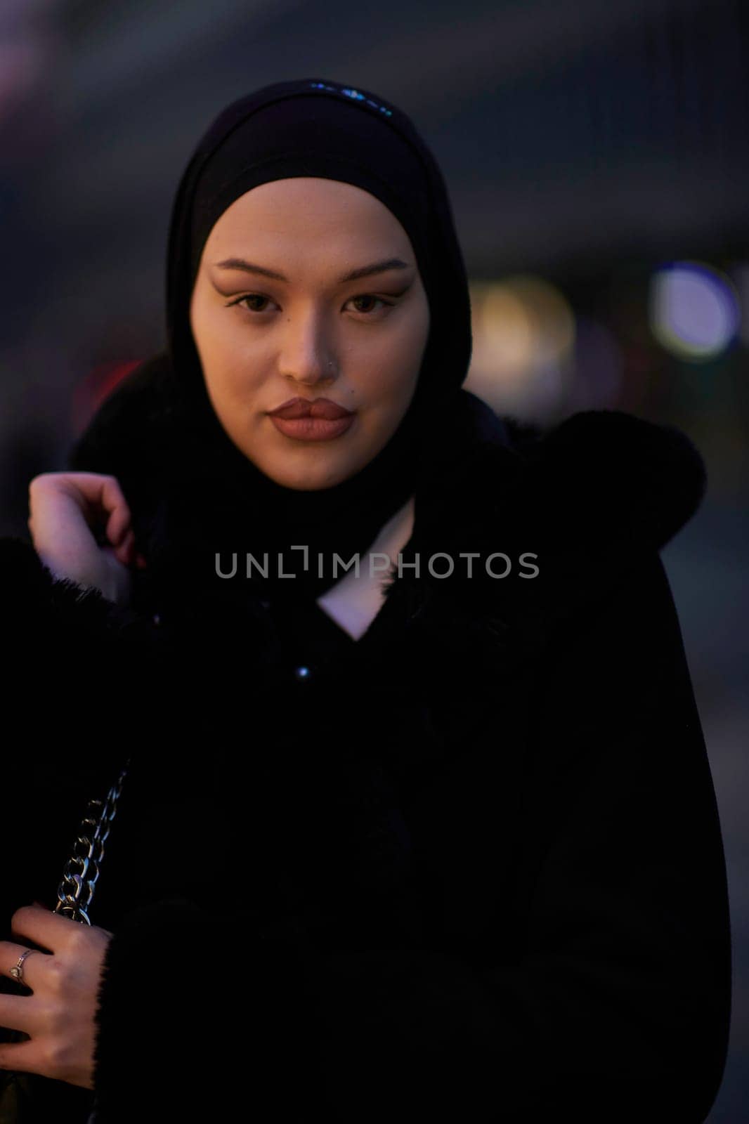 Muslim woman walking on an urban city street on a cold winter night wearing hijab with bokeh city lights in the background