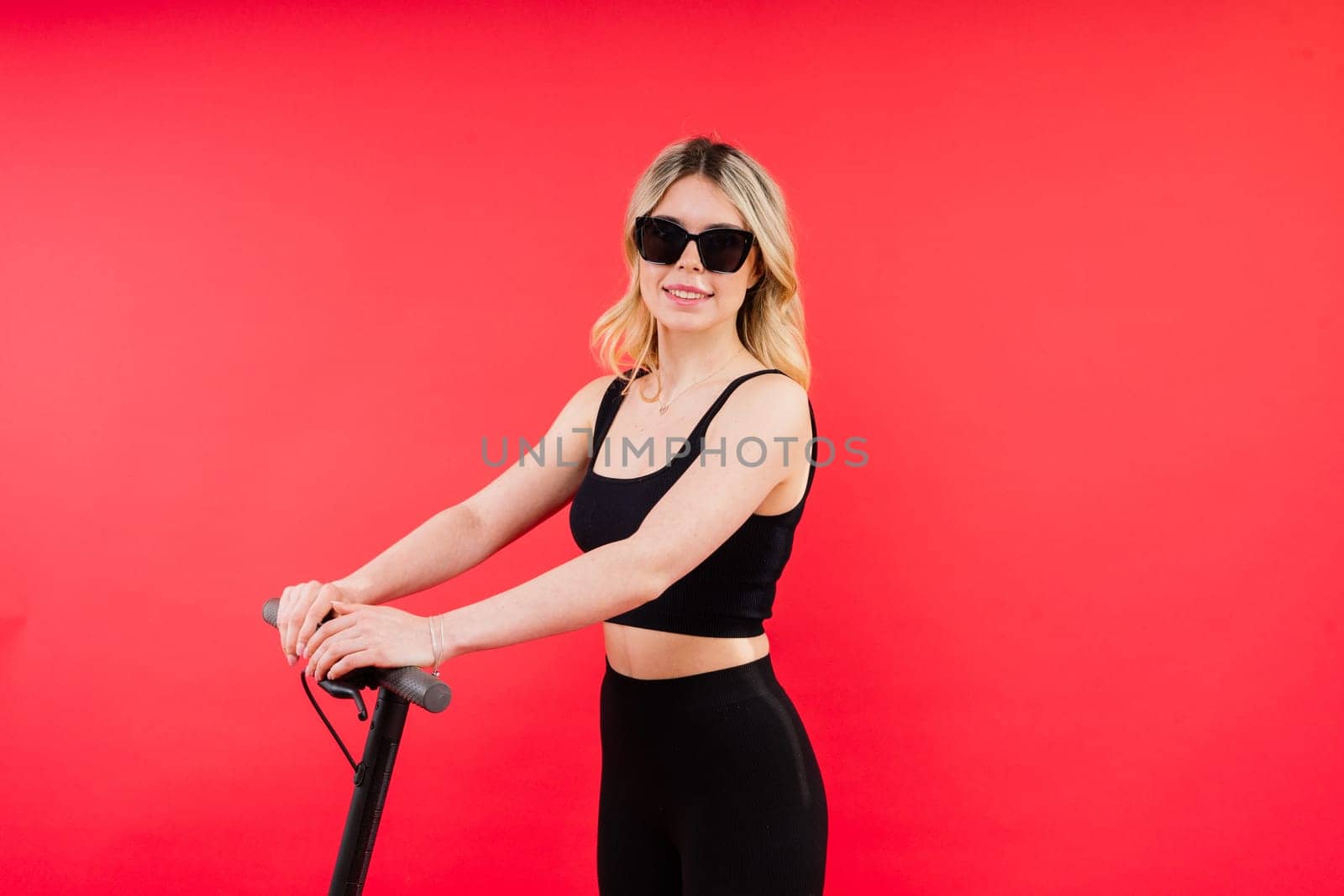 Beautiful emotional young female in a sport clothes on electric scooter on red and white background