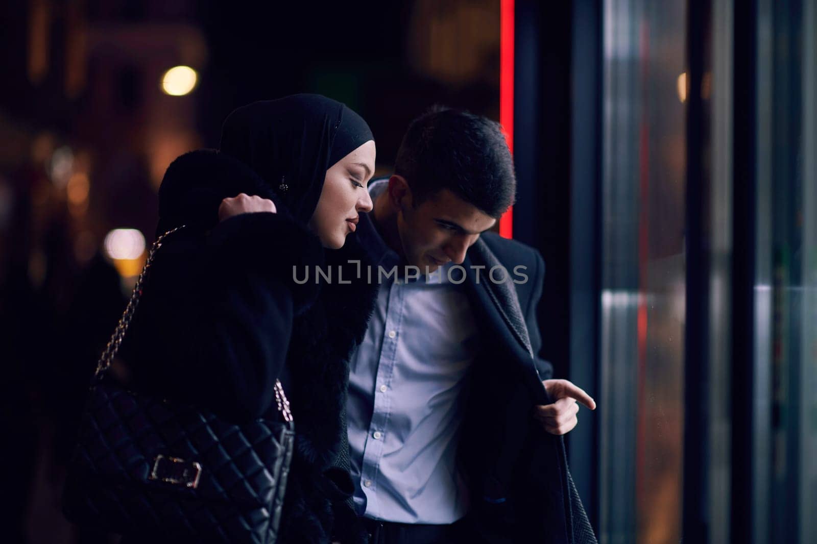 Happy multicultural business couple walking together outdoors in an urban city street at night near a jewelry shopping store window. Successful Arab businessman and European Muslim woman.