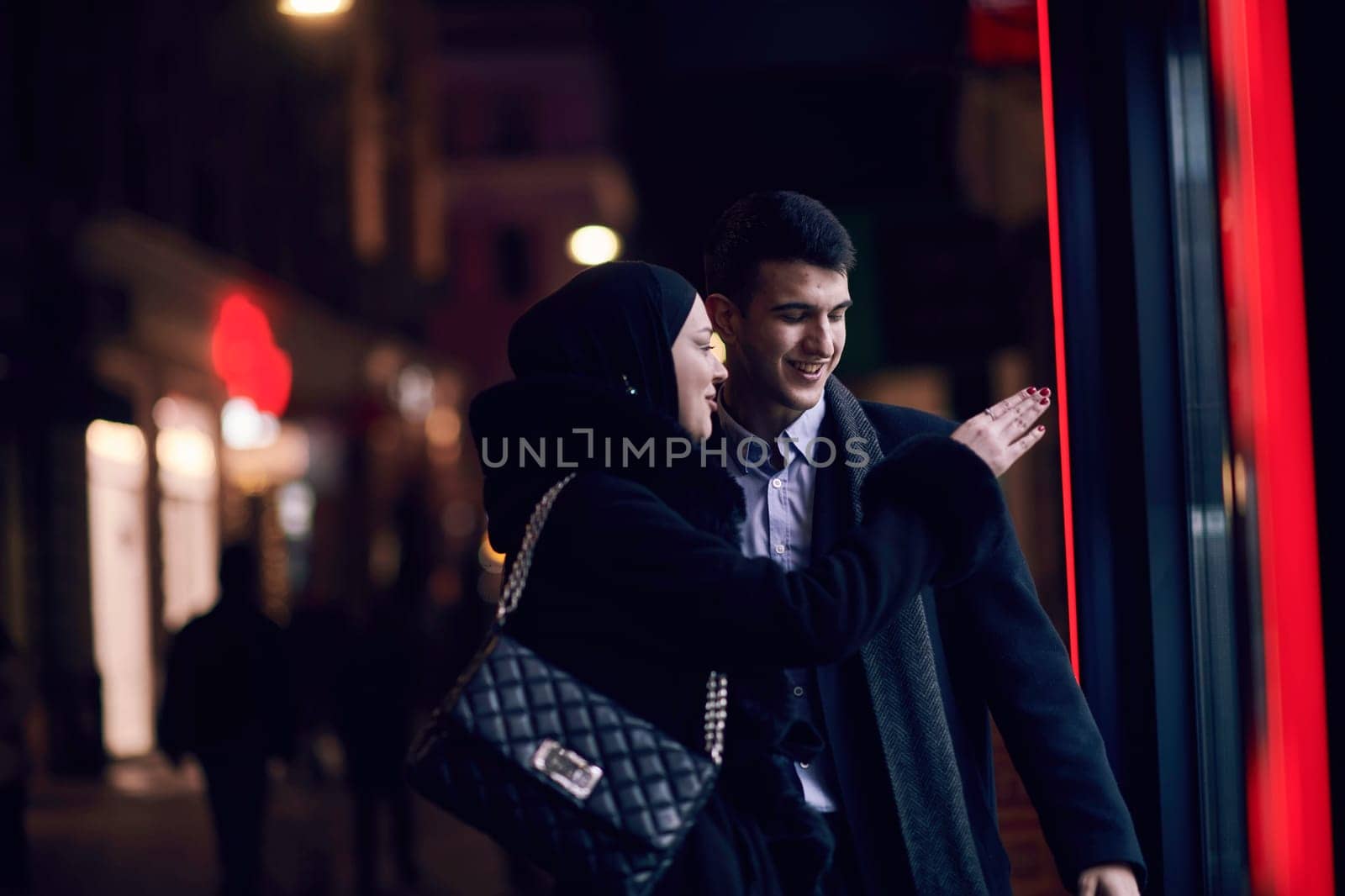 Happy multicultural business couple walking together outdoors in an urban city street at night near a jewelry shopping store window. Successful Arab businessman and European Muslim woman.