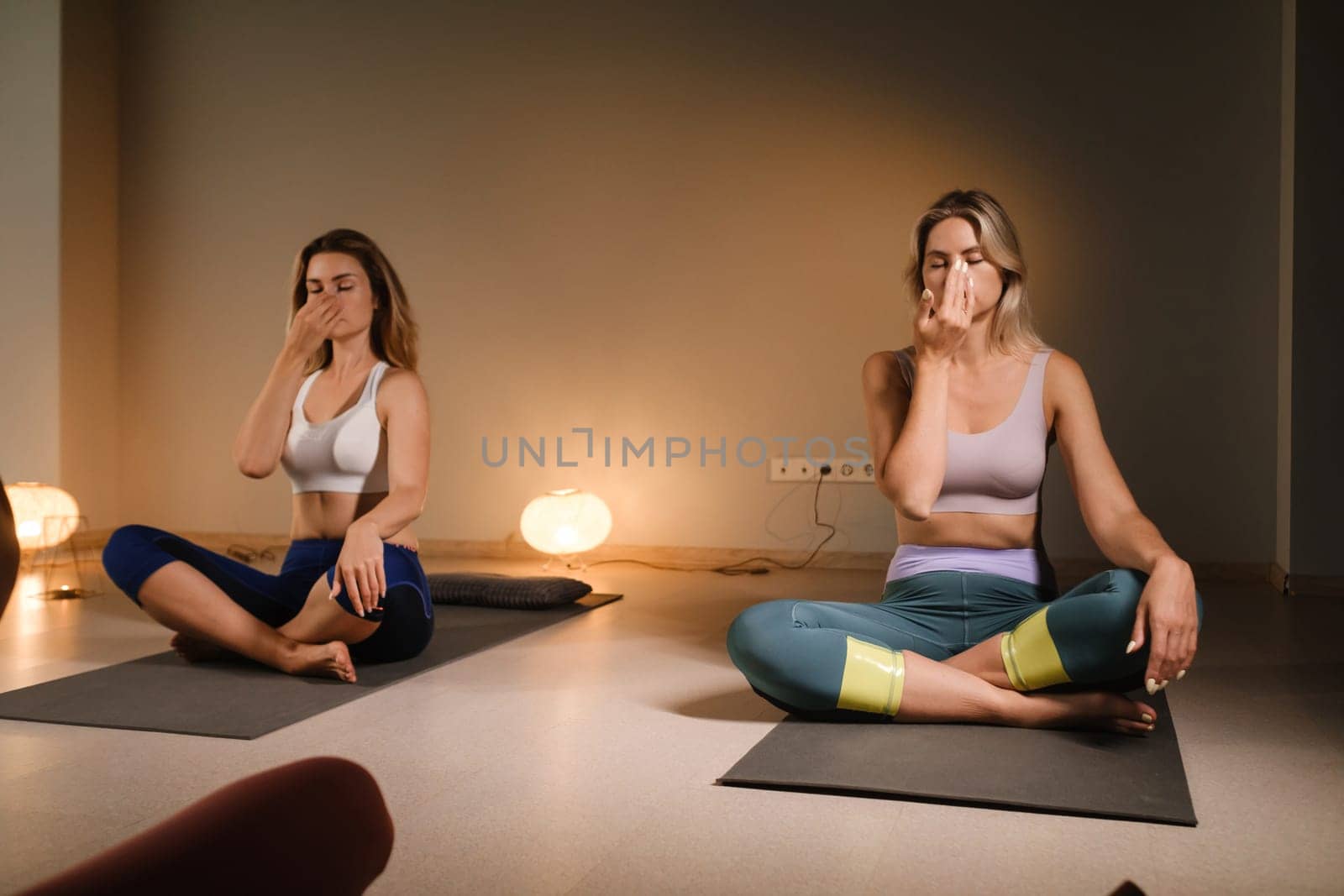 a couple of women performing breathing exercises while sitting on a yoga mat. yoga meditation in the fitness room by Lobachad