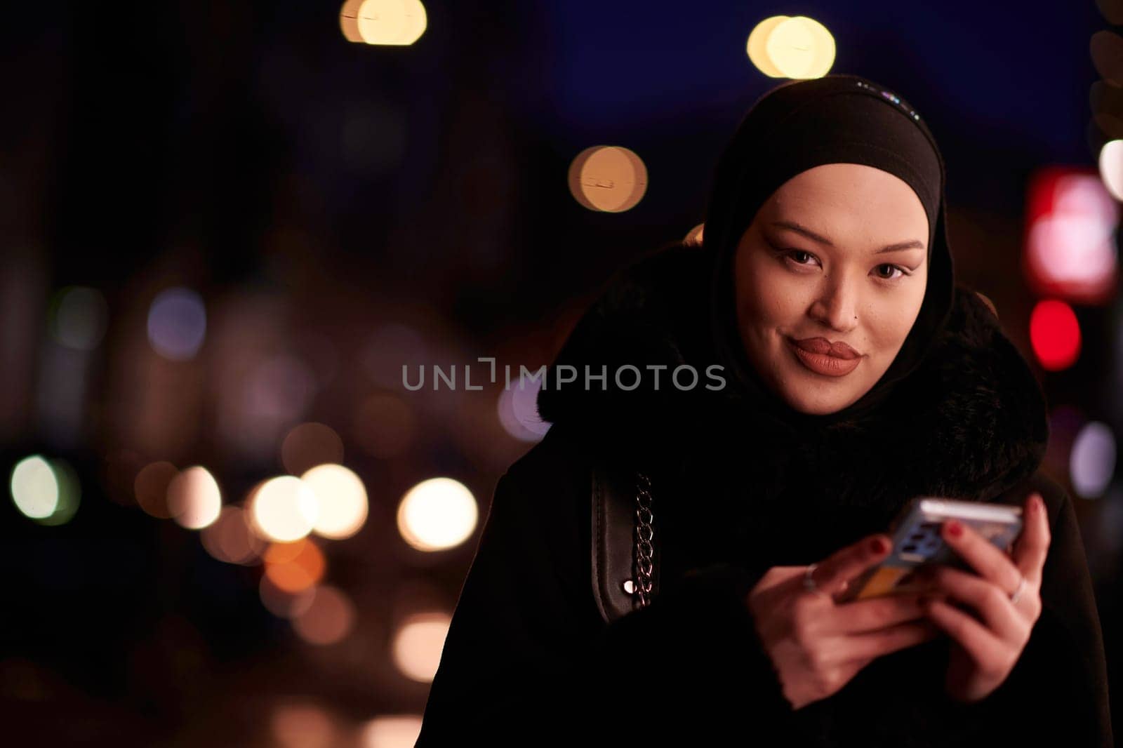 Beautiful and elegant European Muslim Hijabi Business Lady checking her phone and social media on urban city street at night