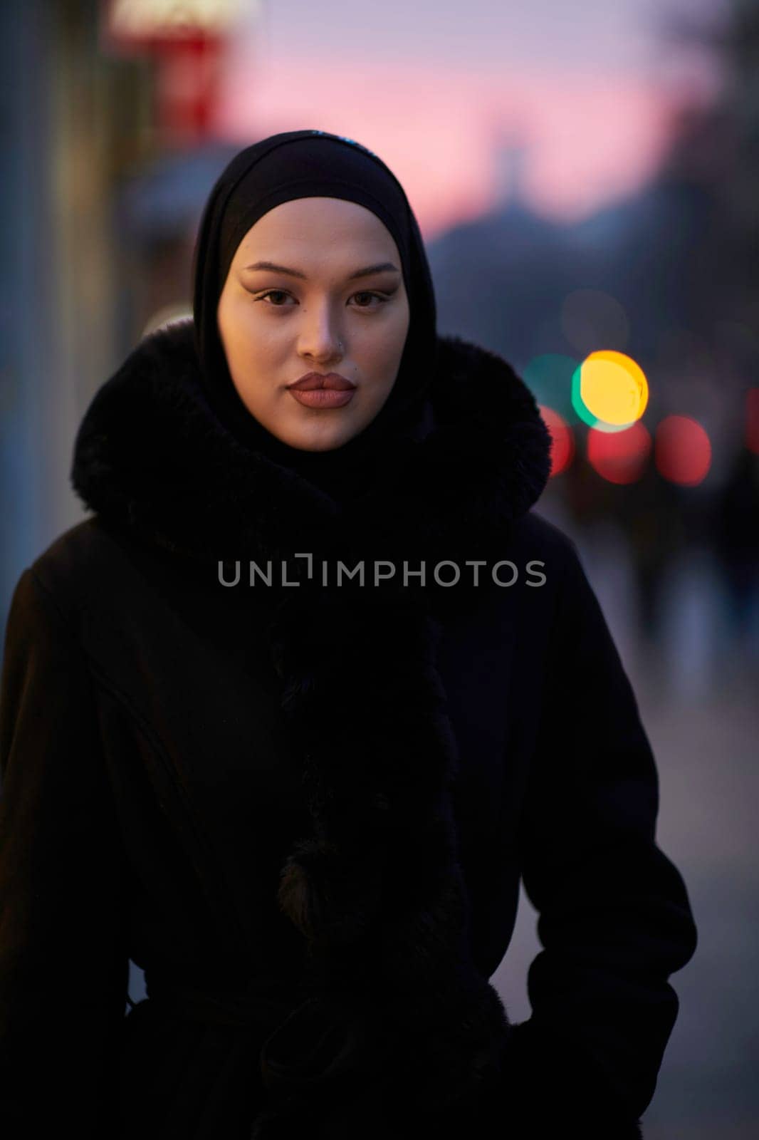 Muslim woman walking on an urban city street on a cold winter night wearing hijab with bokeh city lights in the background