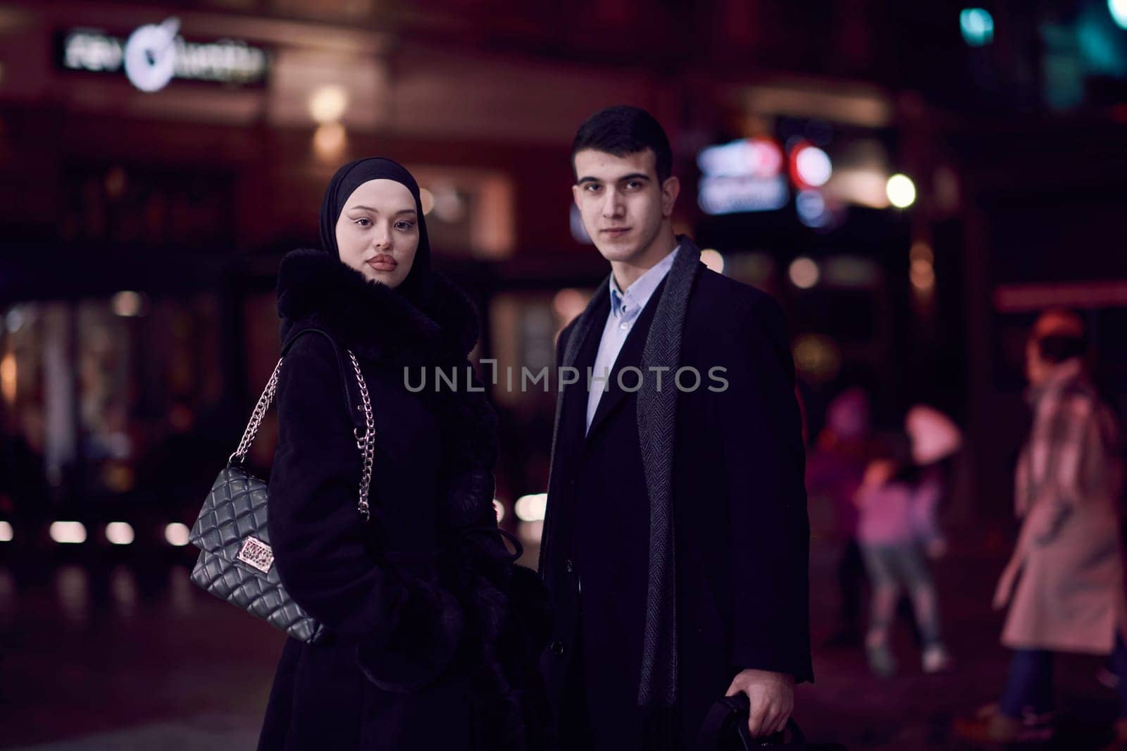 Happy multicultural business couple walking together outdoors in an urban city street at night near a jewelry shopping store window. Successful Arab businessman and European Muslim woman.