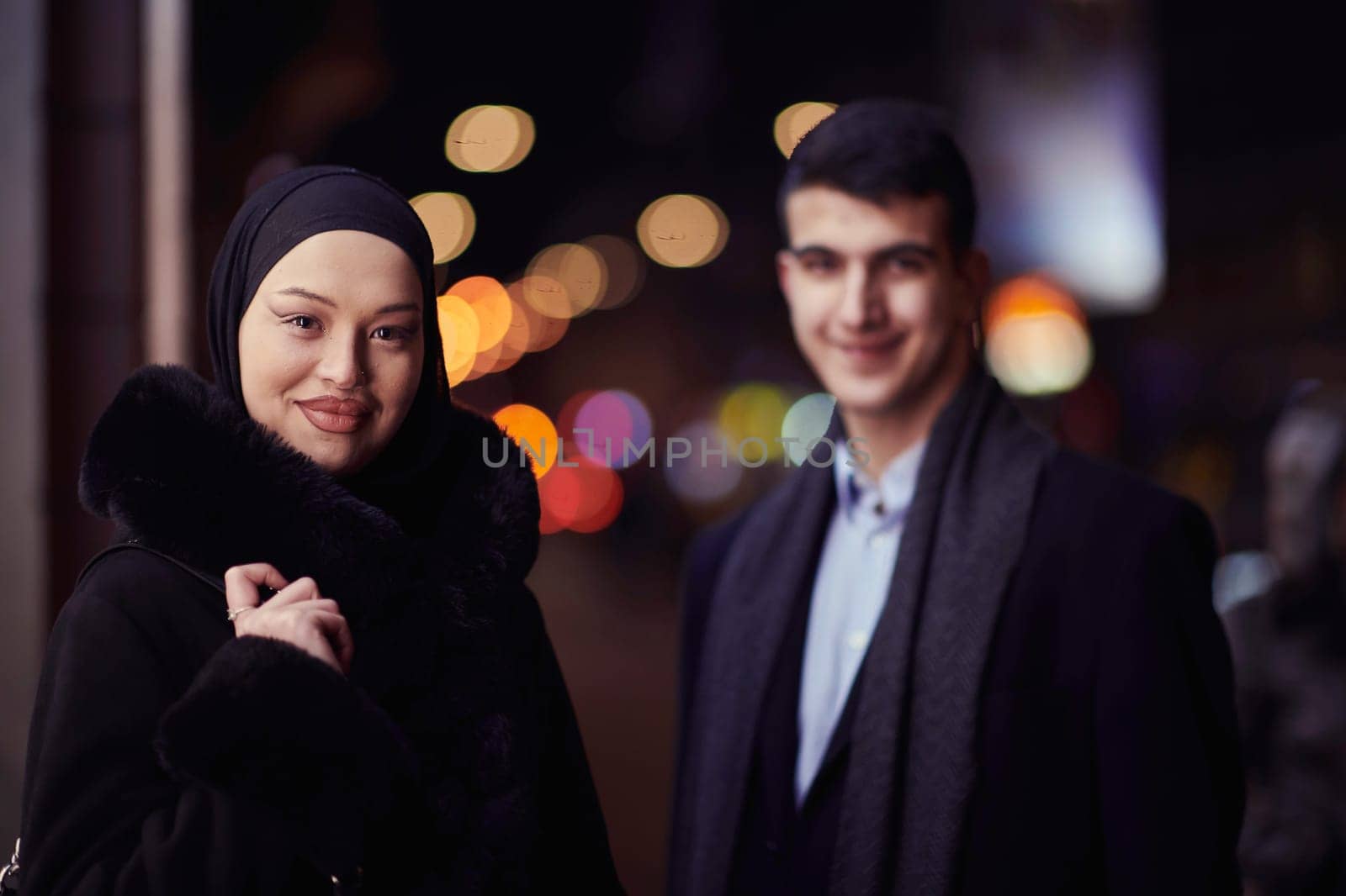 Happy multicultural business couple walking together outdoors in an urban city street at night near a jewelry shopping store window. Successful Arab businessman and European Muslim woman.