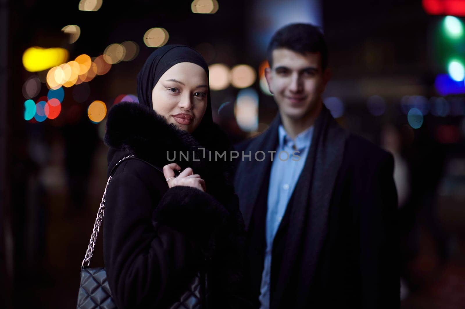 Happy multicultural business couple walking together outdoors in an urban city street at night near a jewelry shopping store window. Successful Arab businessman and European Muslim woman.