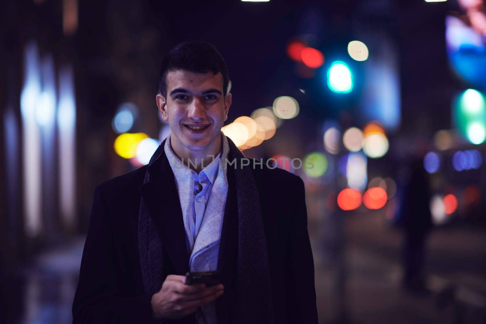 Smiling Meedle Eastern man walking down street near modern office building, freelancer businessman looking away holding mobile phone on busy city street at night