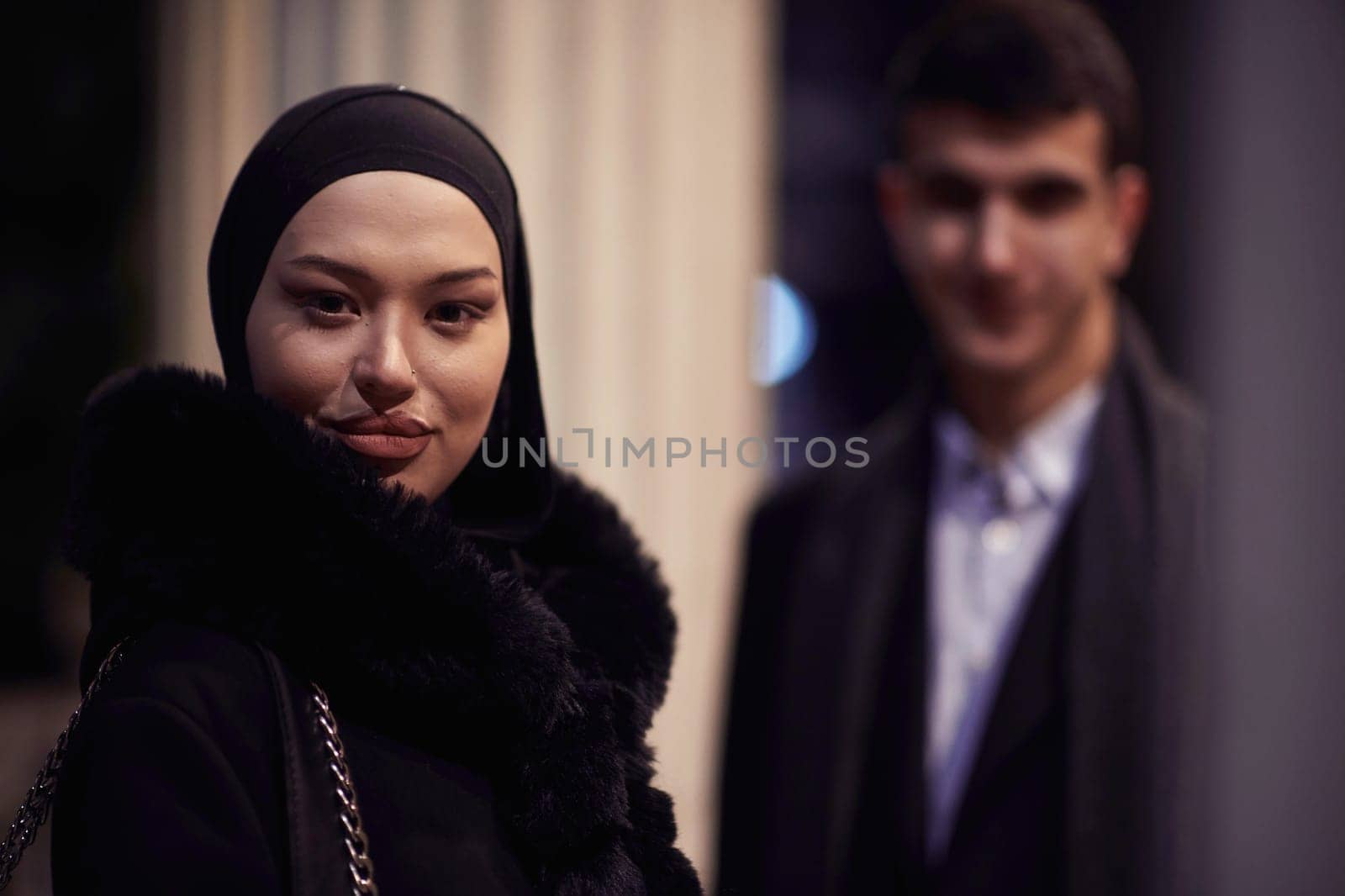 Happy multicultural business couple walking together outdoors in an urban city street at night near a jewelry shopping store window. Successful Arab businessman and European Muslim woman.