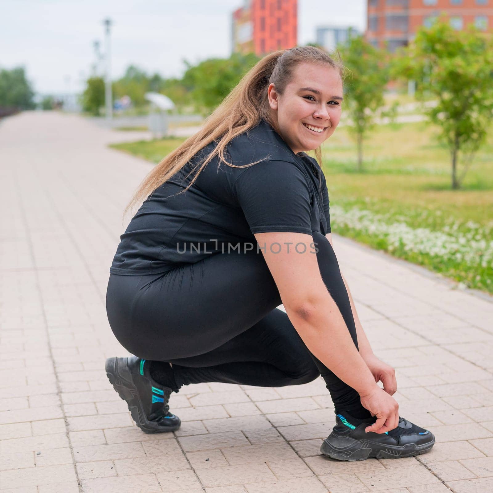 A fat woman in a tracksuit crouches down and ties her shoelaces outdoors