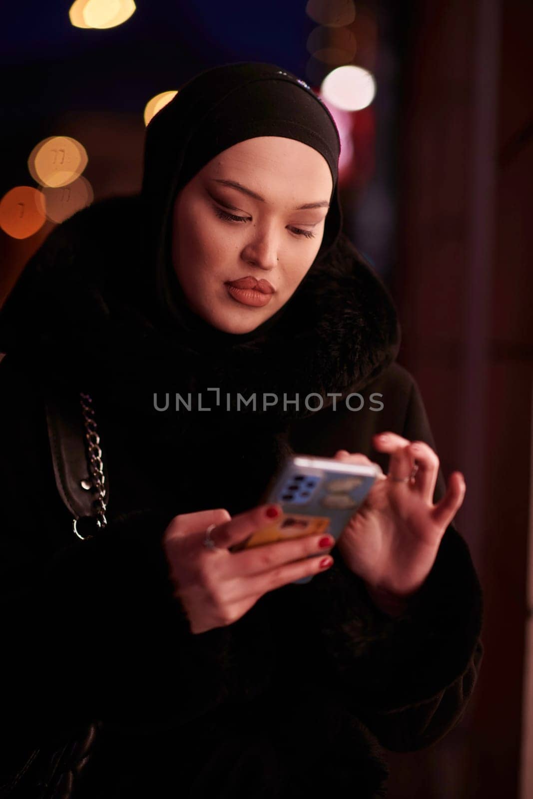 Beautiful and elegant European Muslim Hijabi Business Lady checking her phone and social media on urban city street at night