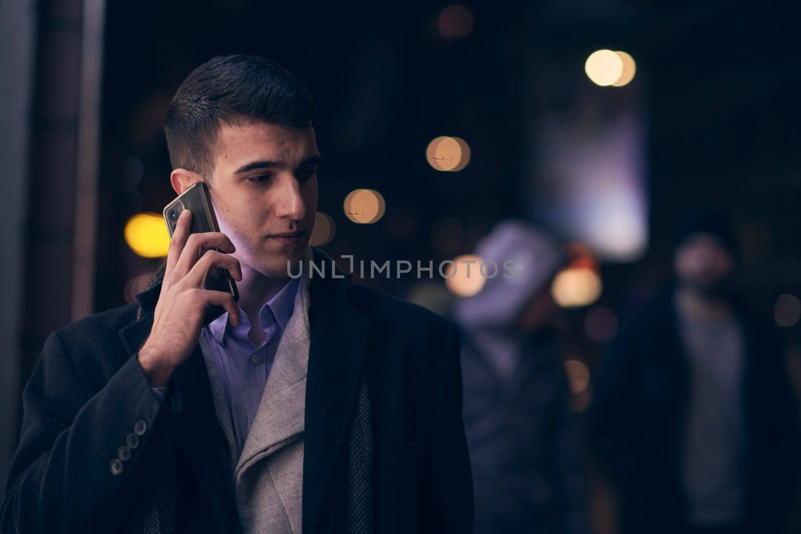 Smiling Meedle Eastern man walking down street near modern office building, freelancer businessman looking away holding mobile phone on busy city street at night