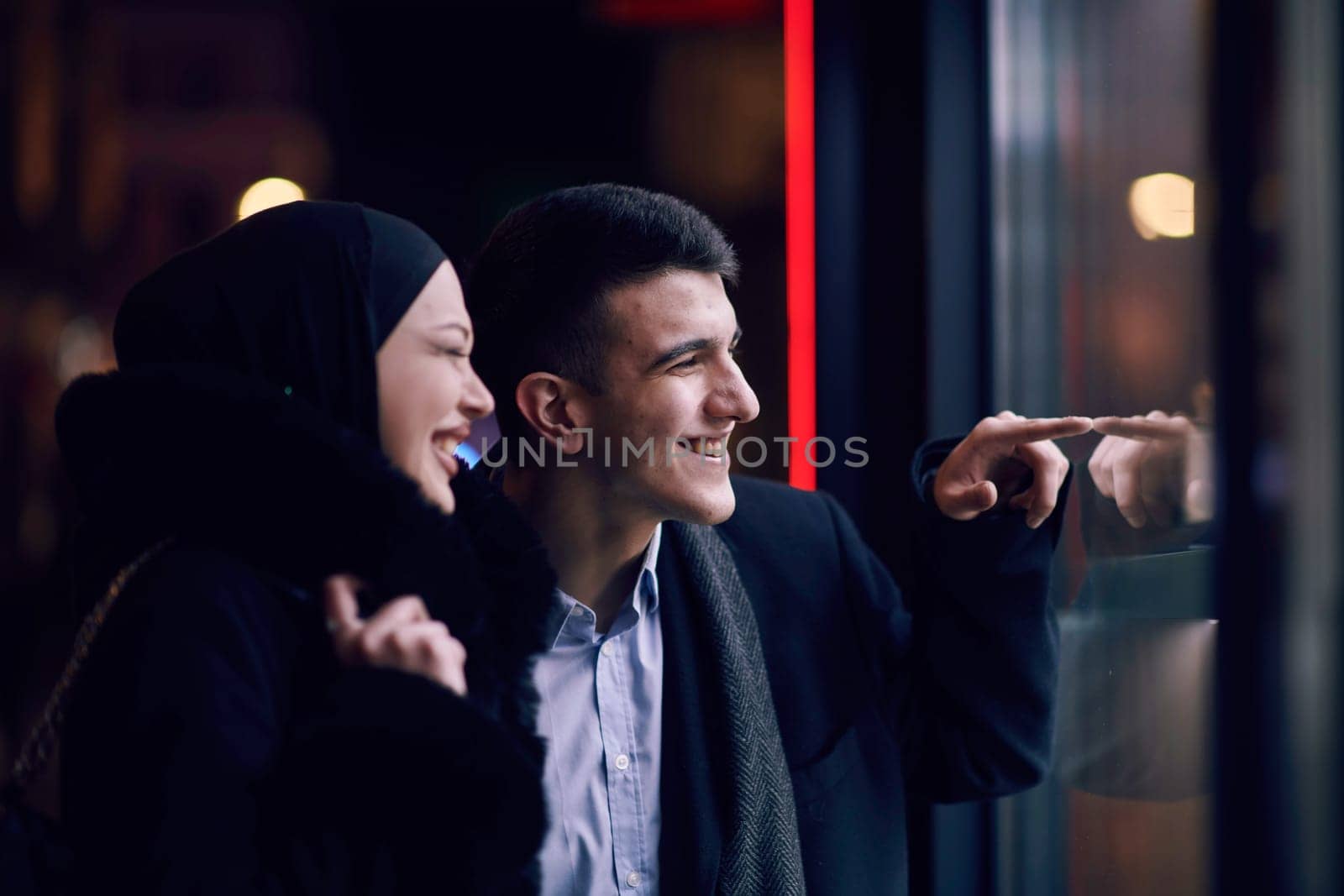 Happy multicultural business couple walking together outdoors in an urban city street at night near a jewelry shopping store window. Successful Arab businessman and European Muslim woman.